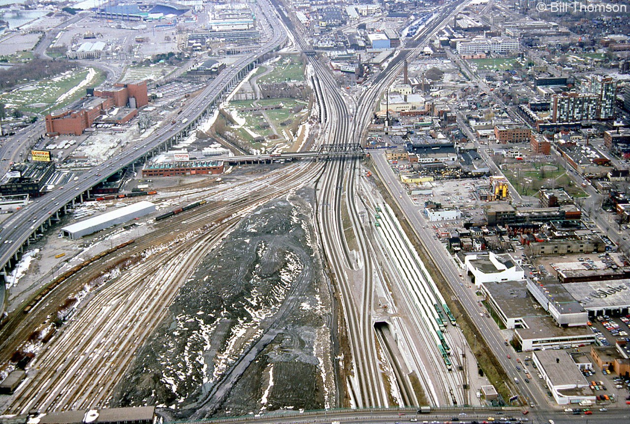 Here's the aerial view of the downtown Toronto railyards, as seen looking west from the CN tower in April 1987. The whole area has and was undergoing many changes: by now many tracks of CN's Spadina Coachyard (bottom left) were either vacant or removed altogether, as VIA had moved their passenger train servicing to the new Toronto Maintenance Centre at Mimico, and all the old downtown railway lands were prime real estate for redevelopment. CN's Spadina Roundhouse (out of frame at the bottom) had also been vacated and would soon be replaced with the Skydome.

In the centre of the image is Bathurst Street bridge where the western rail lines of the Toronto Terminals Railway from Union Station split into the CN Oakville Sub, CN Weston Sub, and CP Galt Sub. The industrial pre-gentrified Liberty Village is visible above the junction (note the bright blue building along Strachan Avenue, part of the old John Inglis factory). Next to it is CP's Parkdale Yard, by then converted into a container-handling facility (full of blue CAST containers).  The Massey Ferguson plants can be seen in the upper right of the photo, a series of manufacturing buildings clustered in the Strachan/King area that once manufactured farm equipment that was shipped out by rail.

On the upper left are the CNE grounds, and below that the Molsons Brewery, a large brown brick building served by rail (accessed via the CP "Wharf Lead" from Parkdale Yard, seen running on an angle across the top of the image above Inglis). Just below is the CBM/St. Mary's Cement Fleet Street plant for local concrete distribution (also served by rail). Along Bathurst Street below the Gardiner Expressway are two warehouses for Loblaws. Equipment from the displaced Canadian Railway Historical Association's former harbourfront museum is visible out back, stored on the spur leading from the old harbourfront trackage that had been removed (included is CN switcher 7988 restored in old orange and black paint, later scrapped).

Along the bottom right of the image is Spadina Avenue, with Front Street at the lower right. "The Globe" lettering on the roof of the old Globe & Mail headquarters is visible (demolished in more recent years). GO Transit commuter trains are stored at the Bathurst North Yard, once a CN freight yard. The CP "shed lead" spur that started under Bathurst St. bridge and ran up to street level at Front & Spadina to access the old freight sheds still appears to be intact.