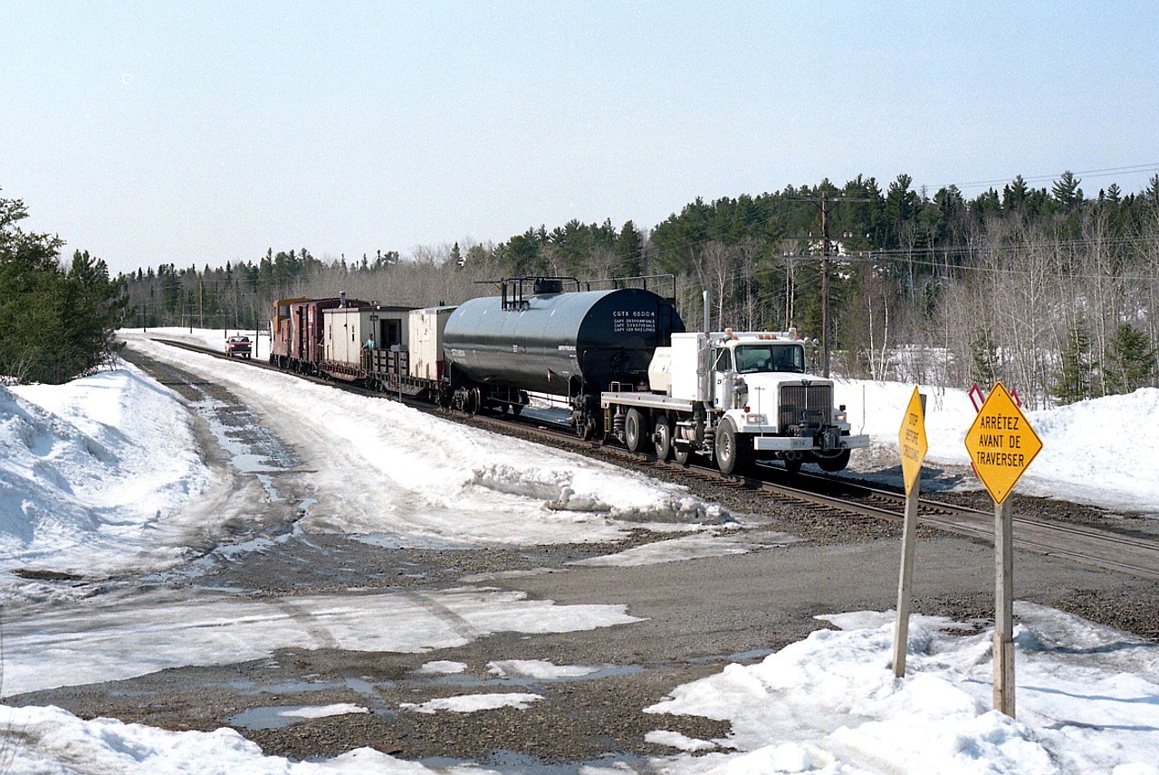 Coming up to the crossing at Geneva Lake Road, 3.3 miles north of Cartier is probably the last Propane Fueling train of the season, topping up tanks. It is early April, and winter is winding down. The train stops and tops up propane at tanks along the way that fuel the Snow Melters which keep switches clean during the harsh Northern Ontario winters. On this train which looks like a Western Star cab in the lead is the huge liquid propane tank car, and behind it a work car which houses the pumping generator as well as perhaps tools and spare parts as needed. An accommodation car is next as this train rolls thru rather remote areas. The boxcar consists of perhaps the main generator and miscellaneous supplies for the train. A caboose is on this run, used as a rider-car or maybe even a Pilot Conductor could be aboard. Following in the hy-rail is the Protecting Foreman, who copies down track protections as well as likely being pressed into service to take the crew to rest as in overnight accommodations if possible.
I'm guessing this train is running Cartier to Chapleau, Stopping to service switch heaters, it must be a rather long and monotonous trip.
Note the two crossing signs. One English, one French.
