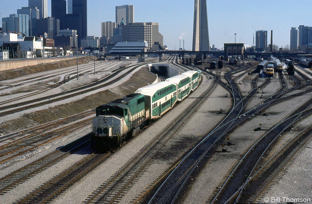 GO Transit GP40-2W 709 trails an inbound commuter train of three bilevels and an APCU (F-unit converted into a cab car/HEP car) heading to Union Station in downtown Toronto, passing by VIA's former CN Spadina coachyard at Bathurst Street. VIA LRC locomotives are visible heading up consists on the yard tracks, and a CN S13 used for switching passenger cars sits in one of the sidings. Self-propelled VIA RDC's sit near the old steam-era coal tower, beyond which is Spadina Roundhouse where VIA's locomotives are serviced.

The 1980's were a transition period for Toronto's old steam-era rail facilities downtown, as the VIA (former CN) Spadina Roundhouse and coachyard would both soon close and move to new facilities in Mimico, and the lands here would be cleared for redevelopment, including for the future Skydome (presently the Rogers Centre). The old CN freight yards at Bathurst Street (on the left) have been converted for storing GO trains, and the new flyunder between Bathurst Street and Spadina Avenue (behind the train) has been completed.