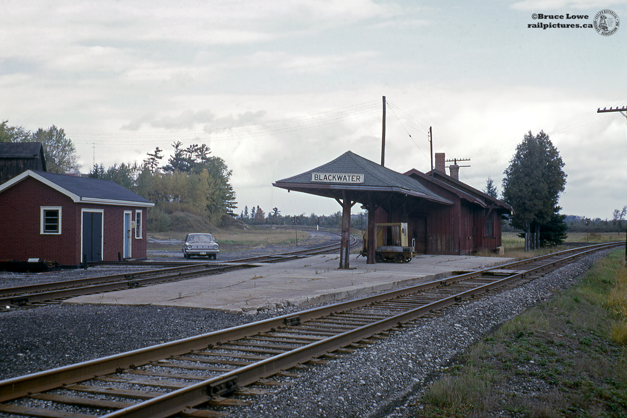 A quiet day at Blackwater Junction finds Bruce and family (in car) making a quick stop by the CNR station, out of use by this time.  The line at right is the Uxbridge sub, mile 18.69, and at left the Coboconk Sub, mile 0.  At left is the section crew's shed for their speeders, both of which are sitting on the station platform.  One older model; open with only front windshield, and one newer enclosed model.  The original station, built by the Toronto and Nipissing Railway in 1871 was named Wick and would be replaced by this structure in 1874 once the Port Whitby & Port Perry Railway reached Wick Junction.  Both merged into the Midland Railway in 1882 before the Grand Trunk would take over in 1884 and eventually Canadian National in 1923.  With the end of passenger service, the station was closed in the early 1960's and demolished about a decade later.Rails through this area began as the narrow gauge Toronto & Nipissing Railway in 1868 headed by George Laidlaw (also in charge of the narrow gauge Toronto, Grey & Bruce Railway), opening from Scarboro Junction; mile 60.56 Uxbridge sub, mile 325.05 Oshawa Sub, to Uxbridge at mile 28.43 in 1871.  The railway would continue building via Wick to Coboconk (mile 36.39 Coboconk sub) halting construction in 1872.  The narrow gauge locomotives powering the line would be constructed by the Avonside Locomotive Works of Bristol, England and shipped across the Atlantic, as well as a small handful built by the Canadian Engine & Machinery Company in Kingston.  Their most unique locomotive was a double-ended 0-6-6-0 wood burner named "Shedden" after the late railway president, John Shedden.  The T&N would further expand with a line from Stouffville to Sutton on Lake Simcoe (reaching Lake Nipissing in North Bay no longer was an achievable goal).  This line opened in 1877 under the name of Lake Simcoe Junction Railway, operated by the T&N to Sutton and Jackson's Point, eventually becoming the CNR's Sutton sub.  The 27-mile Sutton sub would be abandoned by 1930 with only 8.9 miles remaining from the Bala Sub connection at Zephyr to Sutton.  This spur would be lifted in 1981.  In 1882 the T&N, as well as the Port Whitby & Port Perry Railway (PW&PP), Grand Junction Railway of Canada, Belleville & North Hastings Railway, and the Victoria Railway would all be merged into the successful Midland Railway, eventually becoming part of the Grand Trunk in 1884.Mile 0 to 18.69 (Lindsay to Wick/Blackwater Jct) of the Uxbridge sub was completed by the Port Whitby & Port Perry Railway in 1874.  Chartered in 1868, the PW&PP would build north from Whitby to either Port Perry, Port Albert, or Manchester.  Ultimately Port Perry won out with it's location on Lake Scugog and access to the Trent-Severn waterway.  The line was begun in 1869 in the Provincial Broad Gauge of 5 feet, 6 inches to qualify for a premium from the government as avoiding the American standard gauge of 4 feet 8.5 inches kept American railroads at bay, however after a brief bankruptcy and reorganization, the line was converted to standard gauge and finally completed to Port Perry in 1872.  As the railway passed to new owners they felt the need to press north to increase revenues.  Lindsay was selected as the destination; also being served by the Midland Railway (Port Hope-Peterborough-Lindsay-Beaverton) and the Victoria Railway which was under construction as an extension of the Midland Railway from Lindsay to Haliburton.  Extra income from interchange was the PW&PP's goal.  With construction starting to Lindsay the railway changed it's name, first to the Whitby & Port Perry Extension Railway, and finally the Whitby, Port Perry & Lindsay Railway, which would arrive in Lindsay from Port Perry via Manilla in 1877.  From Manilla, a spur was built to Wick to connect with the narrow gauge T&N for more interchange traffic in 1874.  After declining traffic levels, the Whitby, Port Perry & Lindsay Railway would be merged into the Midland Railway along with the few mentioned above in 1882.Today virtually all these branches are history, save for a portion of the Uxbridge Sub used by GO Transit from Scarborough to Lincolnville, and the York Durham Heritage Railway from Stouffville to Uxbridge.  This scene at Blackwater is no more.  The last passenger trains ran in late 1961/early 1962; last listed in the October, 1961 timetable.  The Coboconk sub was shortened to Woodville (mile 12.14) in 1965 before being removed in 1986.  The Uxbridge sub continued in service until 1991 when all track north of Uxbridge was lifted.  Today the line to Lindsay is part of the Trans-Canada Trail while the Coboconk sub is the Beaver River Wetland Trail.More at Blackwater:Blackwater station on Coboconk Sub side in the 1950's.Blackwater Junction in 1958; one train to Lindsay, one train to Coboconk: by Bob Sandusky.Uxbridge Sub side of Blackwater; bob Sandusky, 1958.**Note difference in order boards between both sides of station.Remains of Blackwater with station demolished, Arnold Mooney, 1977.CN gas electric bound for Coboconk.
