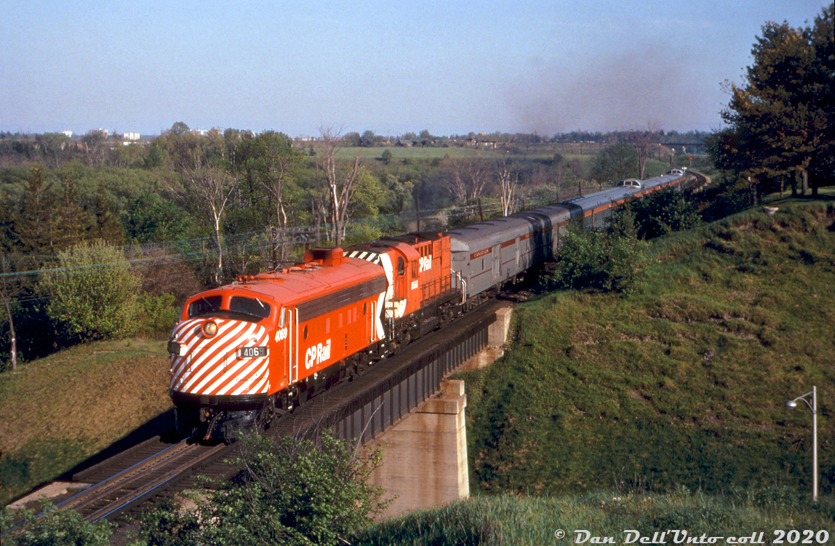A warm spring evening finds freshly painted CP FP7 4069 leading RS10 8568 northbound on Train #11, the Toronto-Sudbury portion of "The Canadian" as they cross over the busy Highway 7 bridge and head north into the town of Woodbridge. #11's previous stop was at West Toronto, but since Woodbridge's station closed in May 1968 the train won't be stopping for passengers again until Alliston. CP's 4066-4075-series dual service freight/passenger FP7 units and steam generator-equipped RS10's were regular power on the Toronto-Sudbury consists at this time. Behind them are two old lightweight steel baggage cars still sporting older maroon letterbands, while the rest of the train are stainless steel Budd cars in modern action red "CP Rail" paint (CP repainted all of its Budd equipment relatively quickly in 1969, but older steel cars often sported maroon letterbands many years after). Note the typical "Skyline" and "Park" dome cars in consist. CP's premier transcontinental passenger train continued to use this routing until VIA took over in 1978 and switched it to running on CN's lines out of Toronto.One can make out CN's Halton Sub in the background (note their high bridge over the Humber River, and the second bridge where the CP MacTier Sub ducked under the line). The train is running on CP's MacTier Subdivision, which was originally the Toronto Grey and Bruce Railway line between Toronto and Bolton (constructed in 1869-1870 as a narrow-gauge line), later coming under control of the Grand Trunk Railway (who standard-gauged the line in 1881) and soon after coming under Canadian Pacific Railway control (via CP's Ontario & Quebec Railway). This part of the line however wasn't original to the TG&B.Time for some local Woodbridge history: the original TG&B alignment through Woodbridge ran through town to the west (branching off to the right where the signal is in the background), but was shifted eastward to this present alignment in 1907 under CP (this involved constructing a new high bridge over the Humber River to the south, and changing the alignment through town including a bridge over Pine Street (today's Woodbridge Ave.)). The old TG&B alignment became a short spur branching south from the station, which lasted into at least the 1980's to serve local industries (there's a segment of rail visible in the parking lot behind Nino D'Aversa Bakery downtown).However, automobiles and horse-drawn carriages continued to cross the CP line here at Highway 7 (then known as MacKenzie Avenue) at-grade, after climbing a winding hill ("Horner's Hill") to the southeast that looped up from the south end of present-day Wallace Avenue to cross the tracks (I believe the old level crossing would have been where the first or second baggage car is today) and continued on what is the present-day stub road MacKenzie Avenue. A large grade separation project undertaken in 1929-1931 saw a new roadway excavated down to run underneath the rail line and the present bridge built, eliminating the jog in the road, the hill and the level crossing. Highway 7 was widened here in 1961, but the old circa 1930 bridge constrains busy Highway 7 to only four lanes of traffic between Kipling and Islington.Charles "Chuck" O. Begg photo, Dan Dell'Unto collection slide.