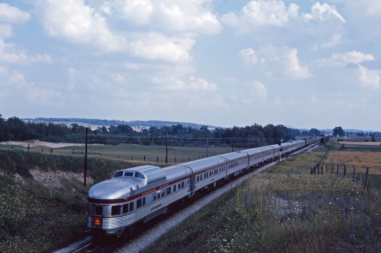 Nearing the end of its transcontinental journey, the train No. 12 The Canadian passes the north switch at the now removed Beeton siding. No. 12, scheduled for a 5:10 PM arrival in Toronto, was one of two daily passenger trains in each direction shown in the summer 1962 time table, the others being The Dominion (train numbers 3 and 4).  The westward Canadian, No. 11, was scheduled to depart Toronto at 5:10 PM. During the summer on Fridays, it was followed 10 minutes later by train No. 317 to MacTier (which returned on Sundays or holiday Mondays as No. 318). The photo was taken from the 9th Line bridge and Jim Brown informs us that the location has now grown in...