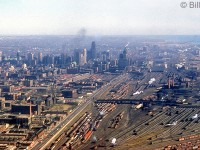 An aerial view of the downtown railyards in Toronto from 1953 yields a very different picture than today. Taken from the air looking east near Bathurst Street, Spadina Avenue is visible crossing over the railway yards. Steam is still dominant, and one can count the white puffs of smoke from many of the steam engines working around the yards. Notable former railway landmarks are CN's Spadina Coachyards, CN's Spadina Roundhouse, CP's John Street Roundhouse, the "High Line" freight bypass running south of both roundhouses (on the right), Union Station, and the CN & CP freight sheds north of Front Street.<br><br>The downtown location of the railway yards was to be in close proxility to busy Union Station, back when train travel was a very prominent and important mode of transportation across the country. Passenger cars would be cleaned, serviced, stocked, and assembled into trains, while the locomotives that pulled them would be coaled, watered and serviced at the roundhouses between runs. The steam era ended on Canadian railways at the end of the 1950's, but the steam era facilities in downtown Toronto continued servicing passenger equipment and diesel locomotives here until they were either closed down or moved to more modern facilities in the 1980's, clearing the way for redevelopment of the old downtown "railway lands" in the following decades.  <br><br><b><i>Some more downtown Toronto photos:</b></i><br>Alternate angle in 1953: <a href=http://www.railpictures.ca/?attachment_id=23318><b>http://www.railpictures.ca/?attachment_id=23318</b></a><br>Looking east from Spadina Avenue: <a href=http://www.railpictures.ca/?attachment_id=28556><b>http://www.railpictures.ca/?attachment_id=28556</b></a><br>Steam at Spadina Roundhouse: <a href=http://www.railpictures.ca/?attachment_id=24444><b>http://www.railpictures.ca/?attachment_id=24444</b></a><br>John Street roundhouse, Union Station & the Royal York Hotel: <a href=http://www.railpictures.ca/?attachment_id=38504><b>http://www.railpictures.ca/?attachment_id=38504</b></a><br>Spadina Roundhouse in 1980: <a href=http://www.railpictures.ca/?attachment_id=32212><b>http://www.railpictures.ca/?attachment_id=32212</b></a><br>Bathurst St. in 1987: <a href=http://www.railpictures.ca/?attachment_id=41906><b>http://www.railpictures.ca/?attachment_id=41906</b></a>