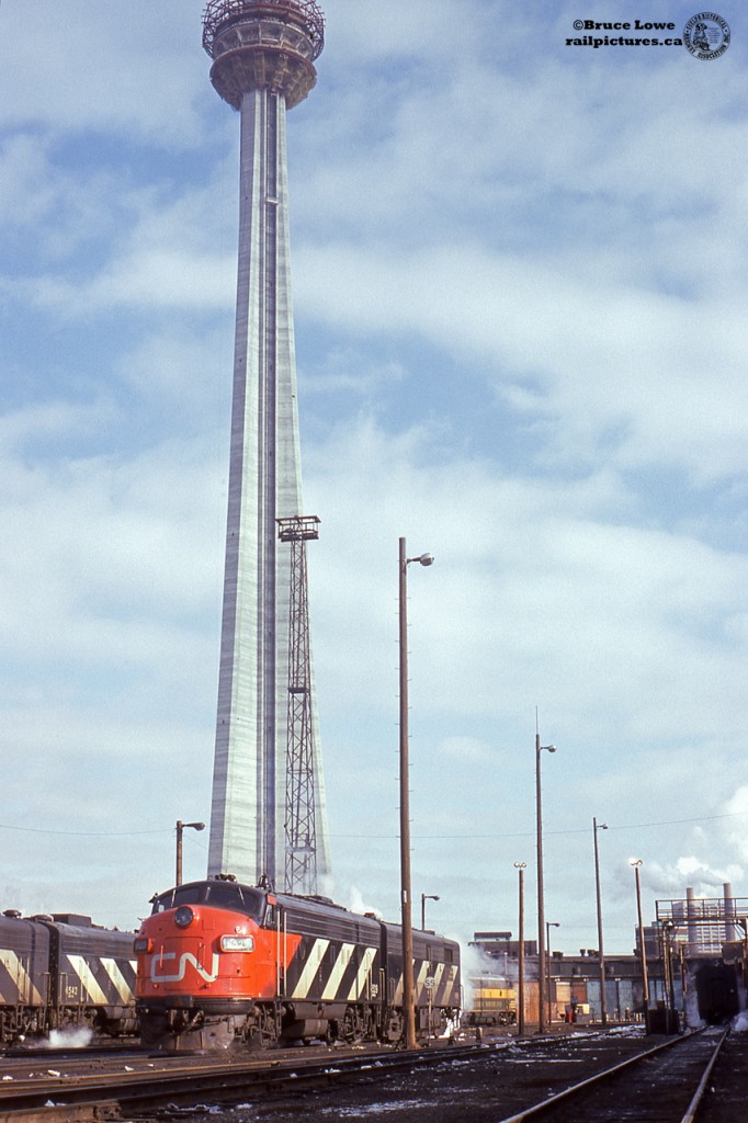Construction is nearing completion on what would be the world's tallest freestanding structure (until 2007).  The CN Tower will open a little over a year later in June 1976.  Meanwhile far below, locomotives idle on the Spadina engine service tracks including CN 6529, (GMD FP9A, 1957) and an unknown MLW FPB4.  At left a pair of GMD cab units, only FP9A 6542 (1958) identifiable, what appears to be the rear of an MLW FPA cab unit at right, and in the distance near the boarded up roundhouse, an ONR FP7A unit.The Spadina roundhouse was built by Canadian National Railways in 1928 and was home to various steam and diesel power off the passenger trains and for local switching duties.CN 6529 and 6542 were transferred to VIA Rail April 1, 1978 at the start of national passenger operations.  6542 would continue in service until 1994 when it was sold to General Scrap in Winnipeg.  6529 was rebuilt by Pointe Ste Charles shops in 1984 and renumbered to VIA 6311, later modified by in 1997 to replace the steam generator with a head end power generator for use out of Winnipeg on the Churchill run.  It would later be sold in 2002 to IFE Leasing to work for the Washington and Idaho Railroad (WIR) as MRLX, later KRLX  6311, wearing the Southern Pacific 2-tone gray 'Lark' scheme, worn by the railroad's Cascade, Lark, and Overland trains.  The unit returned to Canada in 2014 as RPCX 6311, now residing at the Alberta Railway Museum in Edmonton.