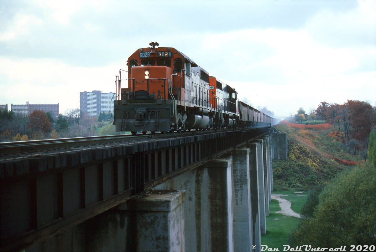 With a cloud of blue smoke kicking up from the brakeshoes of trailing hoppers, CN SD40 units 5029 and 5020 lead an eastbound on the descent downgrade between Rexdale and Weston on the Weston Sub, crossing over the high bridge spanning the Humber River just outside of Weston. This is possibly the "Limehouse Turn", coming from the Indusmin quarry on the Guelph Sub near Acton. The crew didn't get the benefit of dynamic brakes with these units, but timetables state that from Halwest to West Toronto (eastbound) a single 5000-series SD40 was good for 6450 tons. Westbound, factoring in the ruling grade up to Rexdale, the tonnage rating was cut to only 3600 tons per SD40. In the background, one can see some of the apartment buildings off Islington Avenue north of Dixon, and Islington overpass off in the distance. This single-track bridge over the Humber River at Mile 9.4 (formerly "CN McGill") was built by the Grand Trunk Railway in the 1850's when the line was originally constructed as the Brampton Sub (becoming the Weston Sub in 1965). It also spanned over part of the Weston Golf & Country Club's course below, who had a private grade crossing to the west for golf carts. GO Transit purchased the Weston Sub from CN in 2009, and in 2012-2013 the abutments here were expanded and new spans were added to triple-track the bridge for increased GO and UPX passenger train service over the line (click here for a present-day image of the line by Michael Da Costa showing the bridge in the background).Barry Schroeder photo, Dan Dell'Unto slide collection