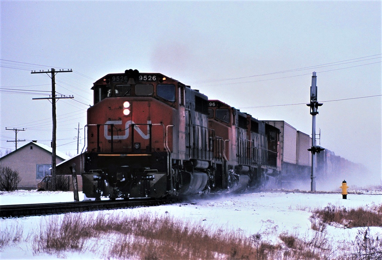On a typical Northern Ontario winter morning, train 221, with typical power for that time period, bounces over Regional Road 80 and the south switch at Hanmer, Ontario at speed.  The short Hanmer siding was used occasionally for storage, was never used as a passing track after CTC was installed, and has since been removed.  Power for the 103 car train was 9526, 9596, and 9537.