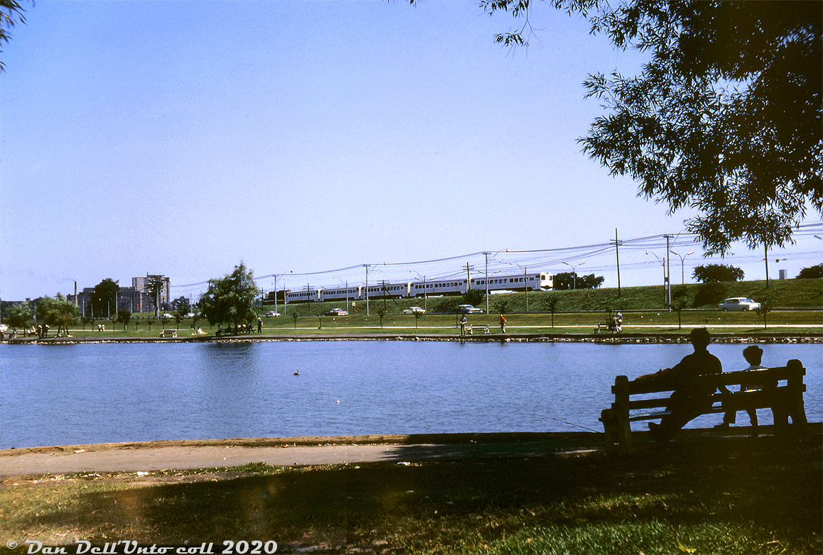 Local residents, including a father and son sitting on a bench with fishing rod set pondside awaiting a bite, enjoy a sunny Summer afternoon at Grenadier Pond in the High Park area of Toronto, as a brand new "GO train" passes by during the first few months of the Government of Ontario's then-experimental GO Transit commuter operation. This area was formerly part of the Village of Swansea, until it was annexed into Toronto earlier in the year. St. Joseph's Hospital in nearby Sunnyside is visible in the distance.

Needless to say, the familiar GO logo and green colours have been a regular sight on trains passing by ever since.

Original photographer unknown, Dan Dell'Unto slide collection