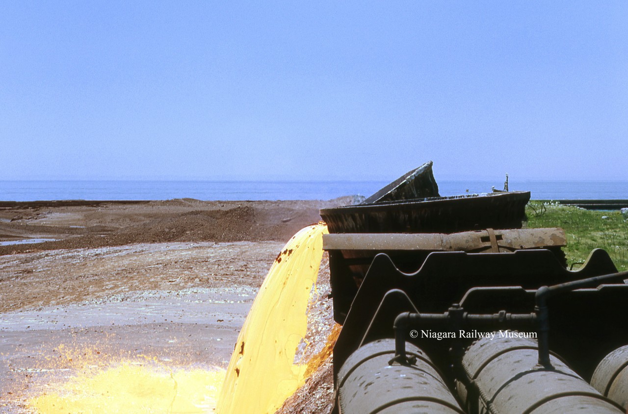 For approximately 65 years from 1912 until 1977 the most visible industrial activity in Port Colborne came from dumping molten slag, the by-product of Iron Making at the Canadian Furnace Division of the Algoma Steel Corporation.  Slag Pots would be shuttled out to the dumping grounds at all hours of the day by GE 50 Ton Center Cab units (#3 and #4) that were ordered in new from GE in the late 1940's.  If the weather was right the operation would light up the entire night sky.  Over the life of the plant the Slag Dump "grew" the East Pier from the foot of Lake Road and Welland Street south into Lake Erie creating an additional 140 acres of land. Today ALLIED MARINE AND INDUSTRIAL has a modern building located where the Blast Furnace was, and the slag dumping grounds are used for ship breaking by INTERNATIONAL MARINE.  The image is looking south, Lake Erie is in the background, the East Break-wall (erected 1880) can be seen on the right.  The Blast Furnace was put on cold idle in March 1977 while the company waited for merchant pig iron pricing to rebound, but by 1981 any hope of that was lost and the Corporation moved to permanently dismantle the factory which was complete by 1982.  By all accounts this was the last dedicated Pig Iron production plant in North America, an end to an era.
