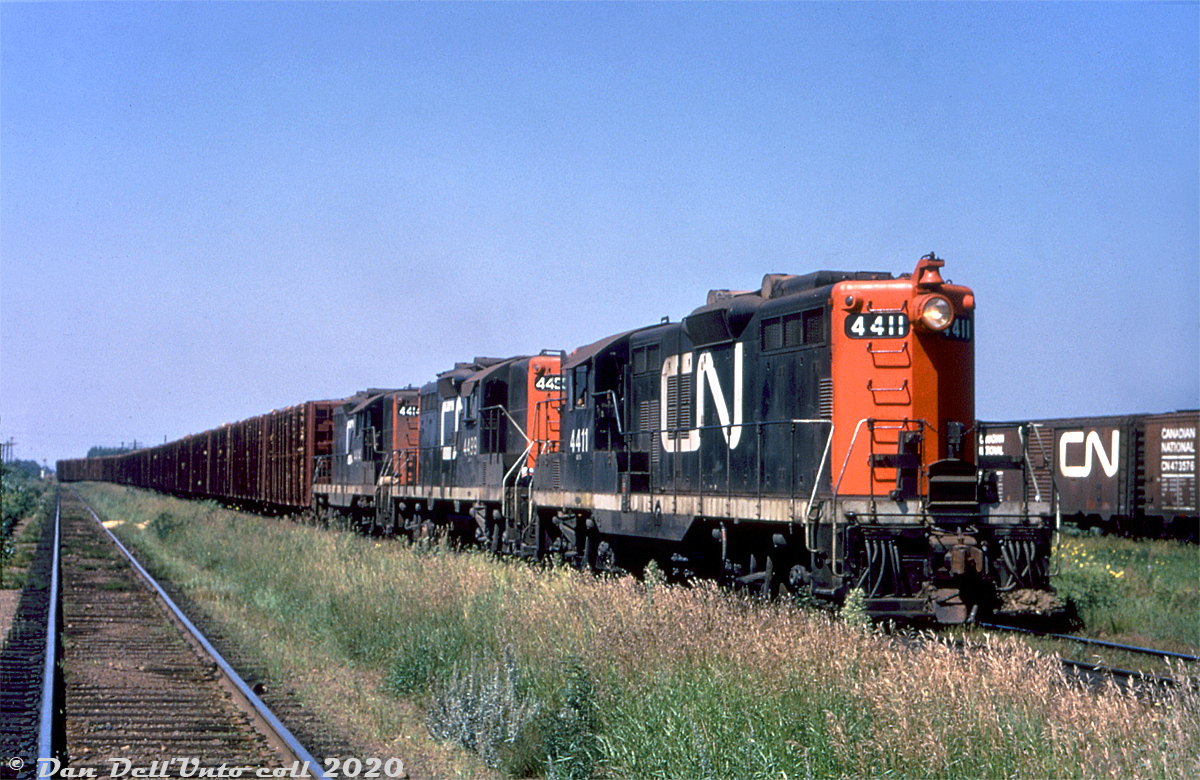 Three CN GP9's, 4411, 4499 & 4414, pull a string of pulpwood racks off the Kashabowie Sub into Neebing Yard in the west end of the Thunder Bay (former Fort William) area, passing a cut of 40' boxcars ubiquitous in these parts for hauling grain to the lakehead elevators. That mound of dirt piled on one of the pilot footboards is curious, maybe the unit hit a small washout somewhere along the line.

The lead and trailings units, 4411 & 4414, part of CN's first GP9 order (4400-4426, built 1955, originally delivered in the 1700-series) and featured the old-style Pyle "garbage can" headlights at both ends. Many later had a dual sealed-beam conversion plate added inside the housing, and eventually the entire headlights were changed out for more modern Pyle dual sealed-beam headlights. Other notable features are the long-hood bell (all of CN's early Geeps were long hood forward), the typical rooftop spark arrestors, early style "bolted" handrail stanchions, and the rectangular brackets welded to the front handrail stanchions (used for mounting CN's early-style removable ditch lights used out west). All three of these GP9's were later cut down and rebuilt as slugs 258, 219 & 234 respectively.

Original photographer unknown (duplicate), Dan Dell'Unto collection slide.