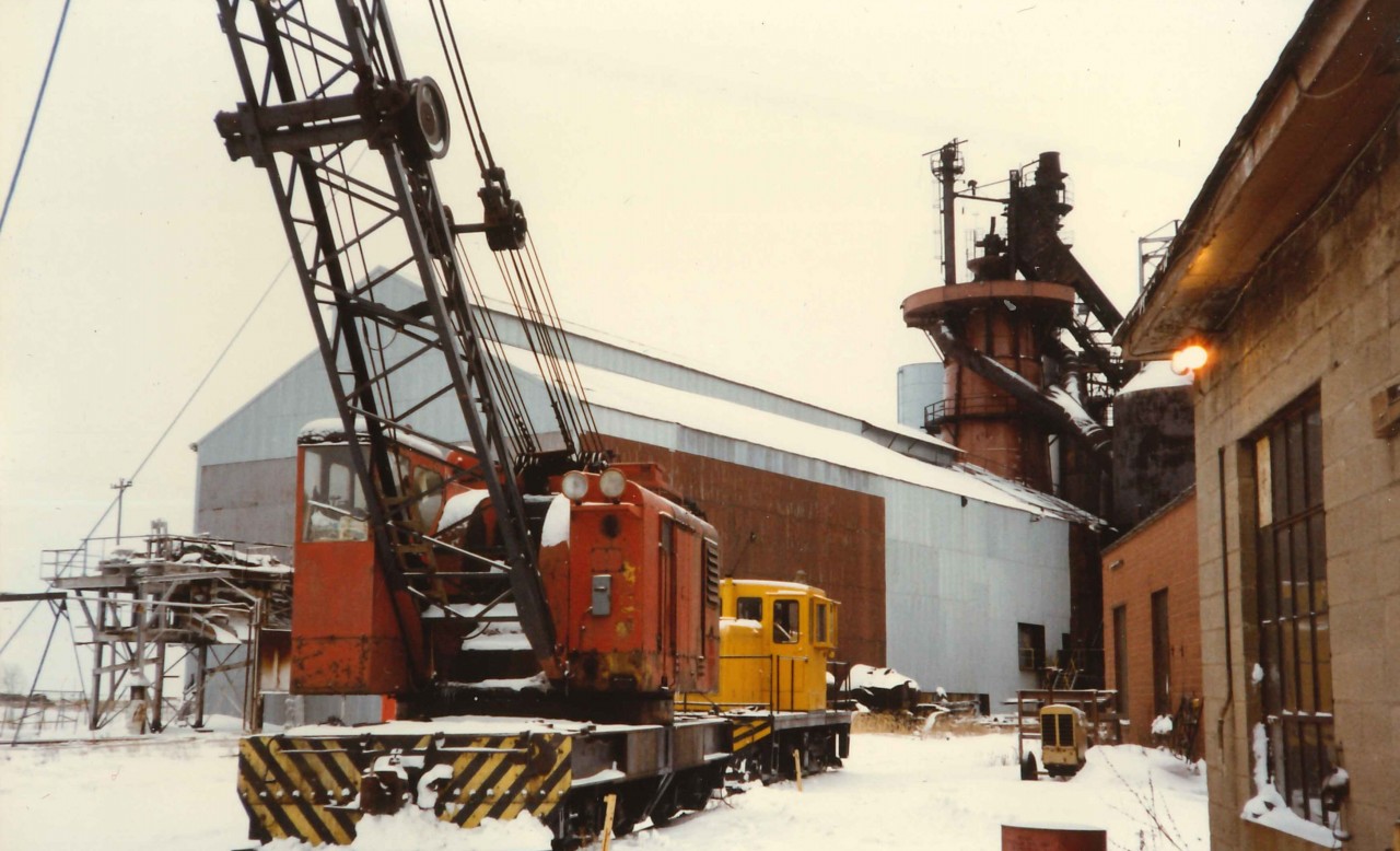 Unnumbered GE 50 Ton Center Cab and American Rail Crane Model 850-80 DE (Serial J3414) sit just north of the Company Engine house waiting their next assignment. In the background the 1912 built Blast Furnace sits as cold as the snow on the ground.  During the Second World War the operation had two (2) Blast Furnaces on site to support the war efforts.  To the left of the Rail Crane is the Pig Casting Machine(exterior to the High Bay), and behind the GE Center Cab are rail bound slag pots tilted on edge. Originally started as the Canadian Furnace Company by American investors from western New York, the operation joined the Algoma Steel Corporation in 1950.  The iron produced at the facility went under the brand name  "Victoria" Pig Iron and was the primary feed stock for iron foundries all over North America.  The image is looking south. Thanks to Paul O'Shell for the specs. on the American Crane.