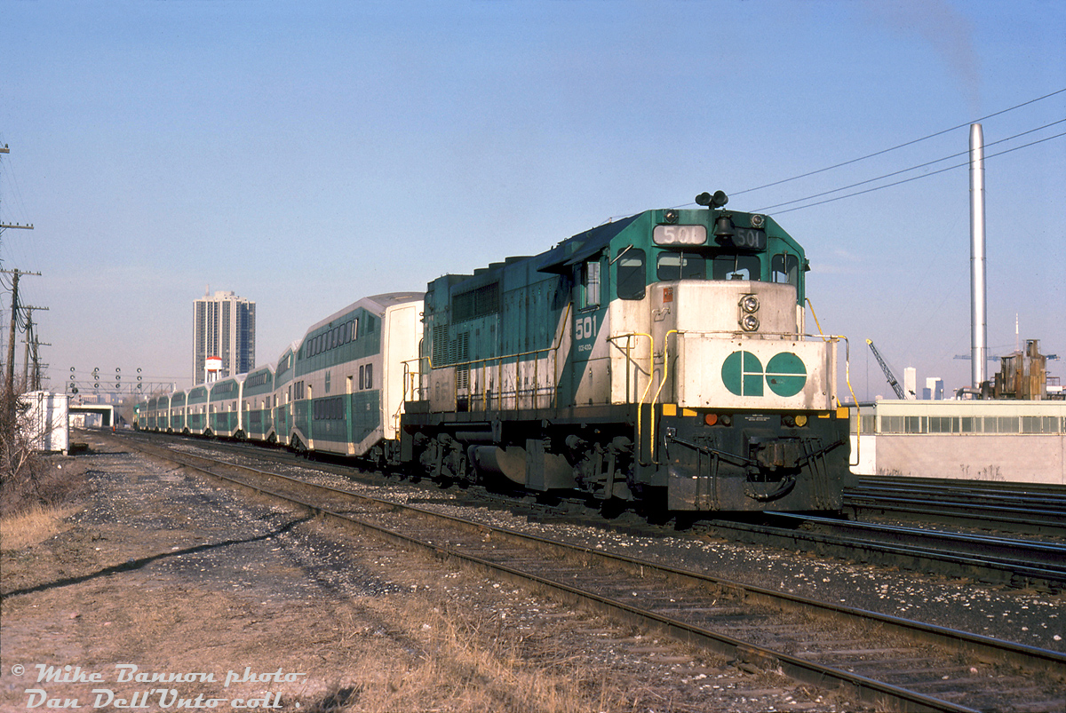 With a GMD GP40TC at either end, a 9-car "L9L" consist with GO 501 on the west end slinks through the crossovers at Mimico East on the Oakville Sub, near Park Lawn Rd. underpass. While this may look like a typical GO consist of the era, the bilevels here were only about 2 years old, all part of the initial order of 80 cars (2000-2079) from Hawker Siddeley Canada in Thunder Bay that began delivery in March 1977. Matching bilevel cab cars wouldn't follow for another few years until the next order in 1983. 

The Queen Elizabeth Expressway overpass is visible in the distance, along with the water tower for the famed Mr. Christie (Christie Brown) factory (opened in 1950, but closed in 2013 and demolished for redevelopment). The apartment tower in the background was real estate developer Bramalea Ltd's new luxury "Palace Pier" tower, completed a year earlier.

Michael Bannon photo, Dan Dell'Unto collection slide.