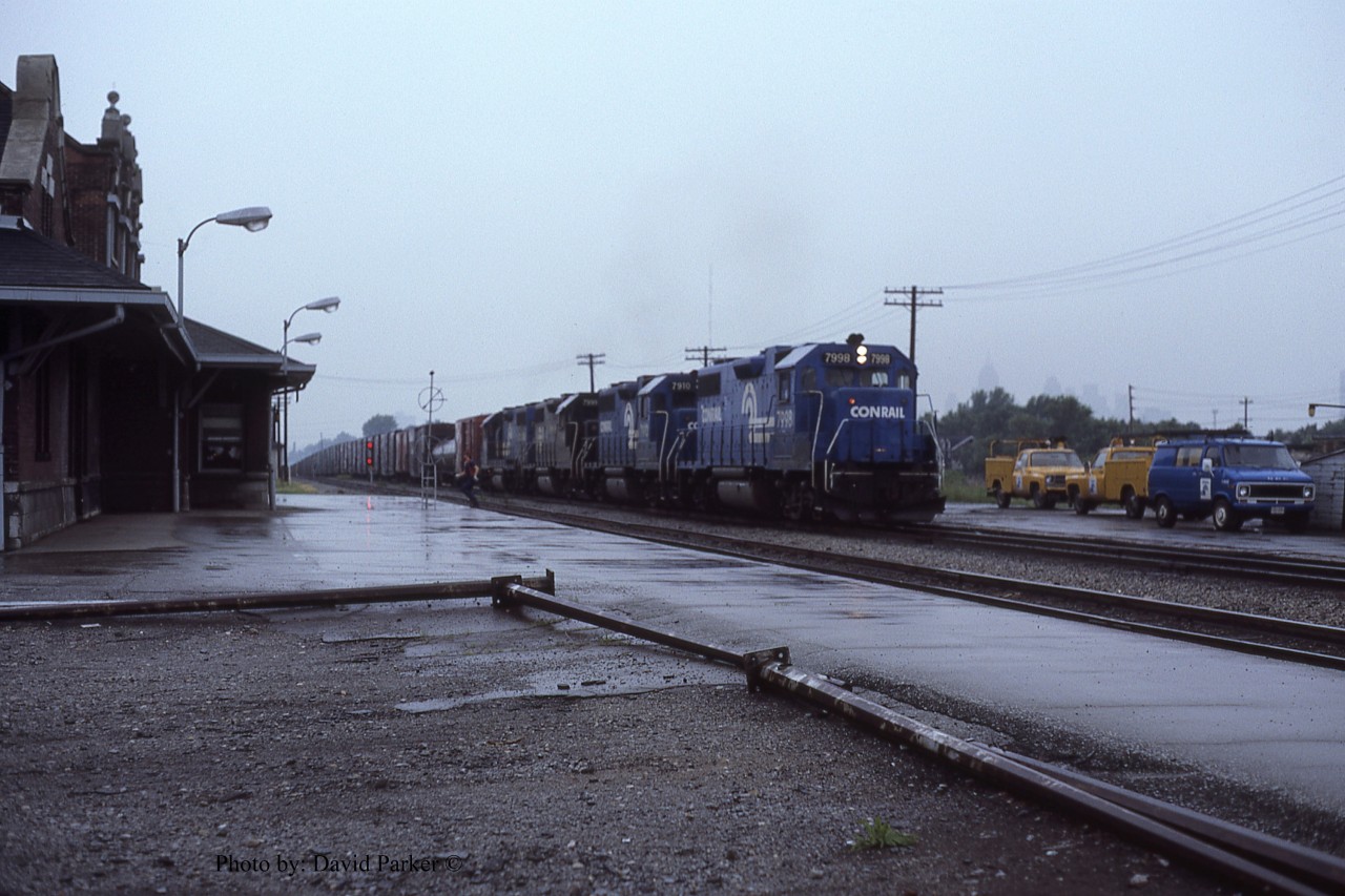 A quartet of Conrail GP38's (7998-7910-7999-7893) claws its way out of the Detroit River Tunnel with train ELDW-3 (Elkhart IN-Detroit/Windsor) and past Windsor Depot on a rainy June 14th 1981. The head end brakeman has bailed off and makes a dash for the shelter of the station where he will wait to make the cut for the Electric Yard. 
  Since the Electric Yard tracks were fairly short, only 45-50 cars, the yardmaster would give the cut number to the operator who would instruct the train, via radio, to stop when the cut was in front of the depot. The brakeman would then make the cut and the power would return after yarding the headend portion to yard the remaining cars. Sometimes a second cut may be necessary.
  ELDW's power would then return 'lite' back to Detroit.