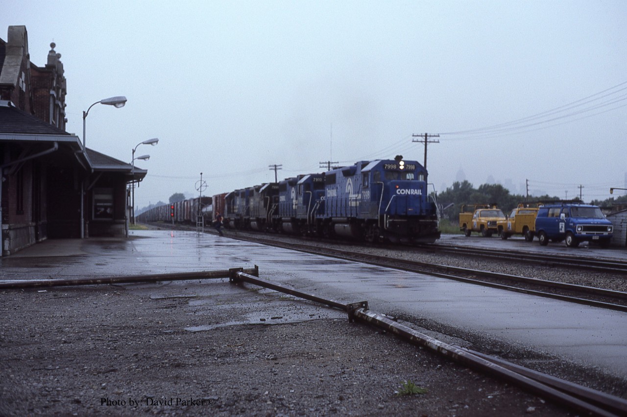 A quartet of Conrail GP38's (7998-7910-7999-7893) claws its way out of the Detroit River Tunnel with train ELDW-3 (Elkhart IN-Detroit/Windsor) and past Windsor Depot on a rainy June 14th 1981. The head end brakeman has bailed off and makes a dash for the shelter of the station where he will wait to make the cut for the Electric Yard. 
  Since the Electric Yard tracks were fairly short, only 45-50 cars, the yardmaster would give the cut number to the operator who would instruct the train, via radio, to stop when the cut was in front of the depot. The brakeman would then make the cut and the power would return after yarding the headend portion to yard the remaining cars. Sometimes a second cut may be necessary.
  ELDW's power would then return 'lite' back to Detroit.