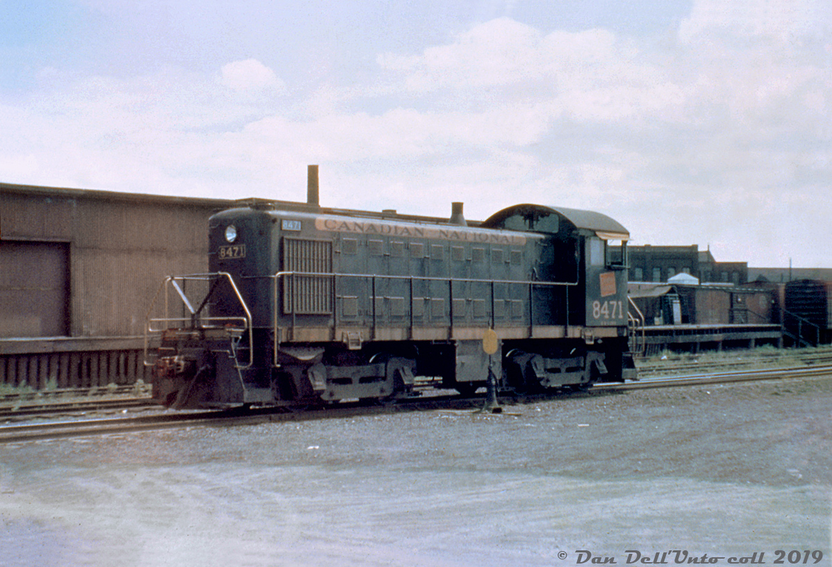CN 8471 (an MLW S3 built in 1953) idles on the service track next to the Parkdale freight shed behind CN's Parkdale Station, just south of the Queen Street underpasses. The two tracks north of the switcher were for spotting cars alongside the freight shed. The boxcars in the background are spotted on one of the two the team tracks by the freight shed. Visible in the background to the right is part of the old American-Abell agricultural manufacturing factory built in 1885 (that later became home to a plate steel manufacturer, then lofts, and eventually demolished 2011 for redevelopment. The nearby Abell Street is a reminder). The freight shed itself became home to a fruit market in later years, and the last remnants were demolished in 2007/08 for the extension of Sudbury Street.

CN and CP both had stations at Parkdale, and even though CP was more well-know in the area for its sprawling Parkdale Yard facilities located on the south side of the rail corridor, CN had a sizable number of industries along the north side of their Brampton (later Weston) Sub to keep their own switchers busy, including the Massey Ferguson lead that ran as streetrunning up Sudbury Street to access the factories north of King Street. Massey Ferguson also had sidings for their buildings on the south side of King around Strachan Ave. And further south were National Casket, the Toronto Refiners, Quality Meat Packers and the Toronto Abbatoir, all north/west of Bathurst Street.

North of Queen there were various forwarding and cartage companies operating out of the Dufferin & Peel area, later including CN's Car-Go-Rail loading ramps, other small industries, and a small storage yard near Lansdowne Avenue where the Newmarket Sub branched off (1961 aerial image here). 

Most of this rail traffic had dried up during the 70's-80's as industry gradually left the old downtown Toronto manufacturing strongholds. CN's Parkdale Station was moved to Sunnyside in 1976 for preservation, but caught fire the next year and was demolished. Many of the old Parkdale industrial lands and factory buildings were still around in the early-mid 2000's (home to lofts, artists spaces, local small businesses, etc), but since then gentrification and rising land prices have seen most redeveloped or replaced by high-rise condos. The rail corridor also retained some of its old character until the late 2000's when the northern portion of Dufferin St. was realigned to connect with its southern half underneath the underpass, and Metrolinx added more tracks and bridge spans for expanded GO and UPX service.

CN 8471 was sold to United Railway Supply (a Quebec-based locomotive dealer and overhaul shop) and renumbered as their 23, but may have been refused by them and ended up at CN's London Reclamation Yard for scrapping in the 80's.

Original photographer unknown, Dan Dell'Unto collection duplicate slide (with a good amount of colour correction and restoration).
