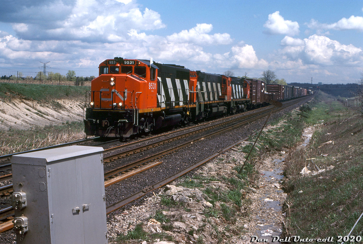One lone white flag clings to the cab of CN GP40-2L(W) 9531 as it, M420 2519, and a 3200-series C424 grind uphill on the grade from the Humber River bridge (CN "Humber", visible in the background but clouded out), operating on a westbound extra freight from MacMillan Yard. The train is just about to cross the grade crossing at Martin Grove Road, denoted by the crossing bungalow on the left. At the time of this photo taken right on the border of Vaughan and Toronto, Steeles still crossed the Halton Sub at-grade, and Martin Grove made a disjointed jog at Steeles.

When CN built the new "Toronto bypass" portion of the Halton Sub in the early-mid 60's, it cut through rural Vaughan and Brampton farmland along the northern reaches of Toronto. Most of the main roads were grade-separated from the start, but a few local roads crossed at-grade including Steeles, Torbram, Airport and The Gore Rd.

But development was moving northward and by the 1980's North York's suburban sprawl was knocking on CN's door. Steeles Avenue had been connected between Martin Grove and Islington and upgraded to become a major east-west thoroughfare along the north edge of the city. Around 1986-87 this area was rebuilt and grade-separated to the current configuration: Martin Grove was re-alinged east to join the southern portion, and meet Steeles at a proper intersection. The CN Halton Sub runs right underneath the intersection of both streets in a box culvert.

Keith Hansen photo, Dan Dell'Unto collection slide.