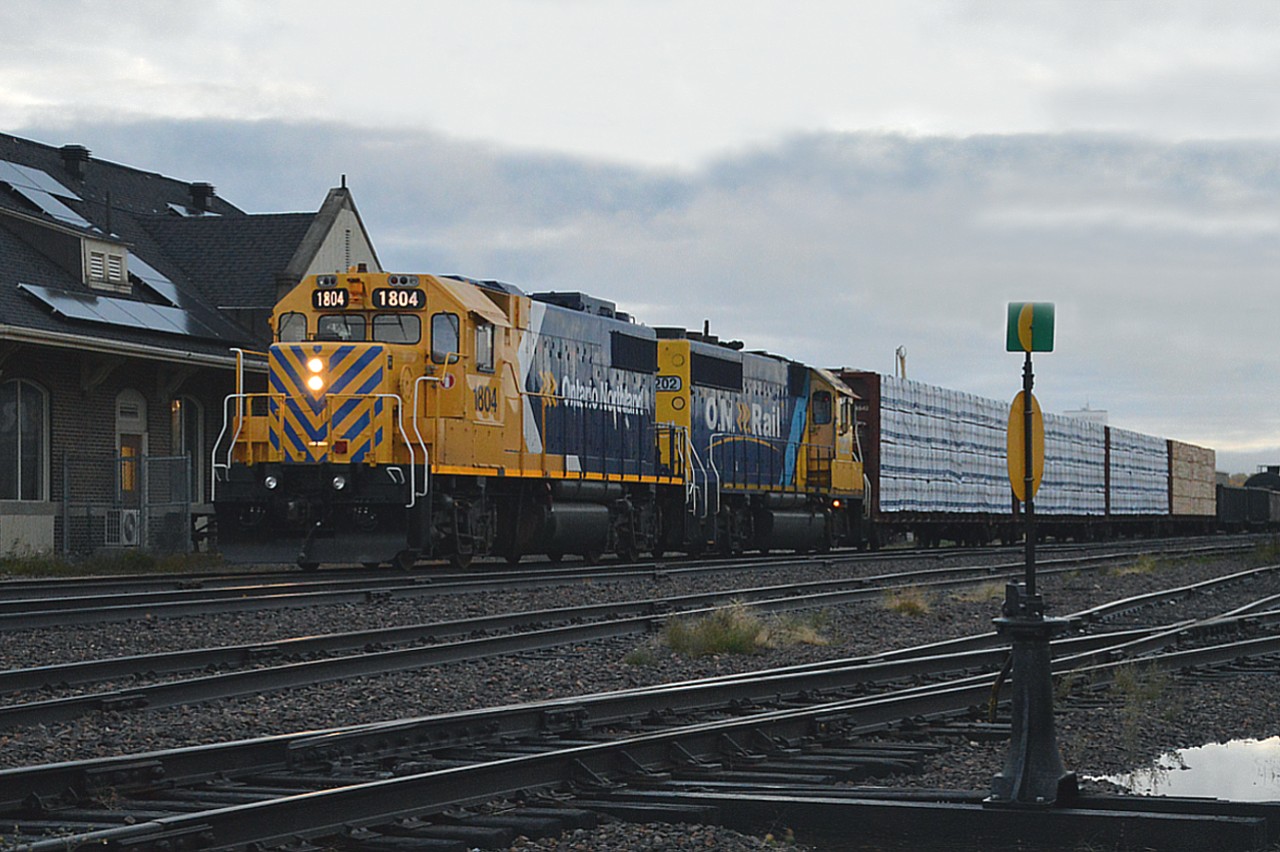 Terrible morning for photography, but what has to be done, has to be done.  We are in a local hotel when the sound of a train horn filled the air. I knew there was some time, as the westward ONR train had to work in town before moving on.
It was just barely daylight. Dark when the train arrived and the sky starting to show some light thru the thick clouds by the time the train was ready to depart after working Rayonier (Tembec) in town. The fact it had been raining actually helped this image.
ONR 1804 and 2202 are just starting to move, past the old (CN) Kapuskasing station (now a museum) on their way to Hearst as I grabbed this shot.  Nice to see two different paint jobs.