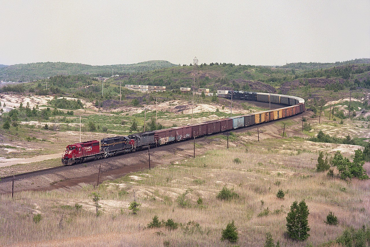 From high up on that slag heap (mine tailings) that overlooks the CP-CN diamond and the town of Coniston; I caught this westbound from North Bay. Great location for train watching.....and best, very few mosquitos.
Scenery is a bit barren but a great view. With the demise of the track from Mattawa down to Ottawa, no major freights can be seen along this line any more. Today this is Ottawa Valley RR (G&W) track.
Power seen is CP 5843, MKCX (Morrison-Knudsen) 9055 and HATX 922. The train is crossing Hwy 17 far in the distance.