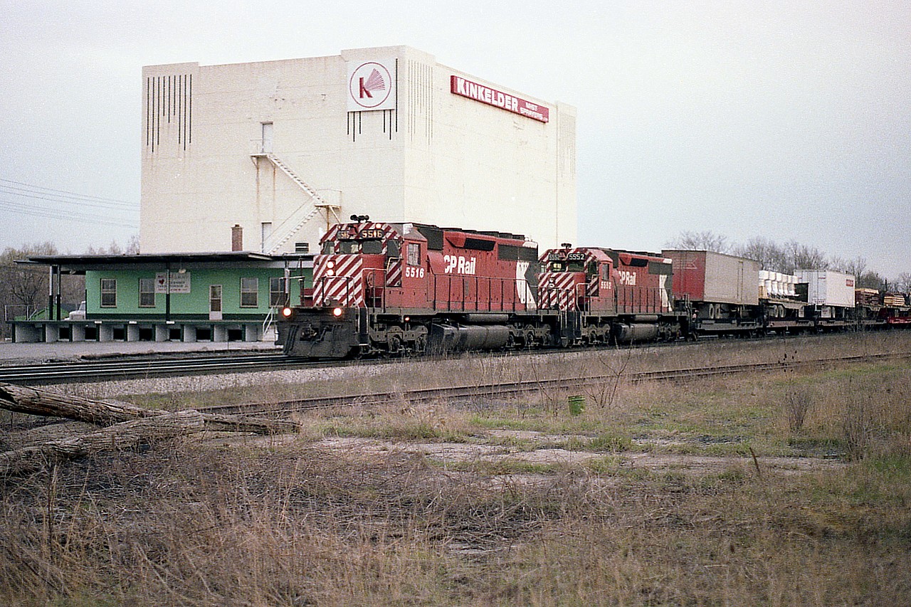 I guess you call this 'mundane times' on the railroad for back in 1980. But what does make this shot of interest is the fact that most viewers on this list under age 30 would have no idea where this was shot. The historical value of photography!!
I am standing where the 403 to Waterdown Rd exit is now. In front of me I believe is the old Waterdown Rd level crossing. The Building is remembered (demo 2009) as 'Aldershot Cold Storage' but for an undetermined amount of time it was home to a branch of Kinkelder Mist Sprayers.  I cannot find any info on that company locating here. They were involved in agricultural sprays which are used on probably mostly fruit trees. The CN main line is still there, but the foreground up to where I am standing is now occupied by the two track lead to Aldershot Yard. Throw in the fact this is a version of the TH&B "Starlite' with CP 5516 and 5552 heading east, and you have what is now an interesting scene. This train always featured 'extra' flags, and was known for its unconventional assortment of freight.