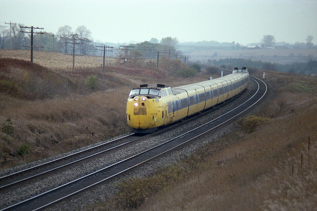 A bunch of us fans gathered on a rather miserable cold day; alternating bits of sun but mostly cloudy and damp; to catch the last run of the rather infamous aluminum bodied CN Turbo Train, heading east to Montreal. The location we chose was a wooden bridge over the line in Newtonville, popular among the local fans. I have forgotten the exact location, but it apparently became known for a time as the "Bee Bridge". An explanation for this can be found in the comment section of RP photo #6832.
The gas turbine powered Turbo, built by United Aircraft, was a bit of a lemon from the start, with various problems sidelining it. On the first trial run in Dec 1968, it hit a truck at a crossing near Kingston. Not a good omen. Five sets were originally ordered, and one of those caught fire and burned the control car and a couple of coaches near Morrisburg in 1975.
Due to various conditions, the train was kept to a 95 MPH limit, although it was capable of much faster speeds.
I had this in mind when shooting this image. Using film, dull days meant slower shutter speeds, and the blurring of such a fast train was very likely; so shot it quite a ways before it got to where we were standing.
Anyway. This is history. None of the trainsets were preserved, and I have no idea when or where this one was scrapped.  Anybody??