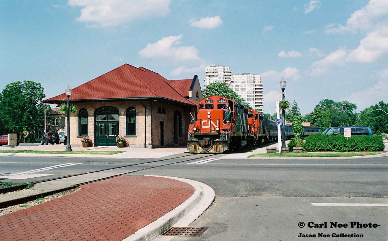 During summer 1997 the railway scene in Waterloo, Ontario was buzzin with activity. The long awaited Waterloo-St. Jacobs Railway (WSJR) had finally arrived and the newly constructed station near Erb and Caroline Streets had been completed and was ready to go. The former Les Trains Touristiques du St-Laurent had arrived that June complete with two FP9u’s along with several passenger cars. Following the bankruptcy of Les Trains Touristiques du St-Laurent, WSJR had purchased all the equipment at an auction. The future was bright and the Waterloo Spur to Elmira was being given an injection of new life with FP9’s now powering long-awaited tourist trains between Waterloo and St. Jacobs much like a retro throw-back to the CN passenger excursions that had powered Sugar Bush Express excursions to Elmira during the 1970’s. 

While the WSJR was now busy operating on the spur, CN continued to provide regular freight service to industries on the line at Waterloo, St. Jacobs and Elmira that were all still frequently receiving cars. While both WSJR and CN operated separately, there were rare occasions when the two interchanged equipment. One instance was the highly anticipated move of former VIA Rail coaches that WSJR had also purchased to operate with their newly acquired equipment from Quebec. 

Here in the sweltering heat of a summer afternoon, CN GP9RM’s 4134 and 4137 are seen about to cross Regina Street in Uptown Waterloo as they slowly haul nine-ex VIA Rail coaches for WSJR as well as their regular freight traffic for Elmira, which required the use of two units on this day’s CN 15:30 Job.