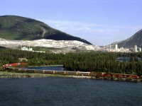 Westbound autos/containers train 115 passes Gap Lake in the Bow River valley. LaFarge's Exshaw cement plant lies behind.