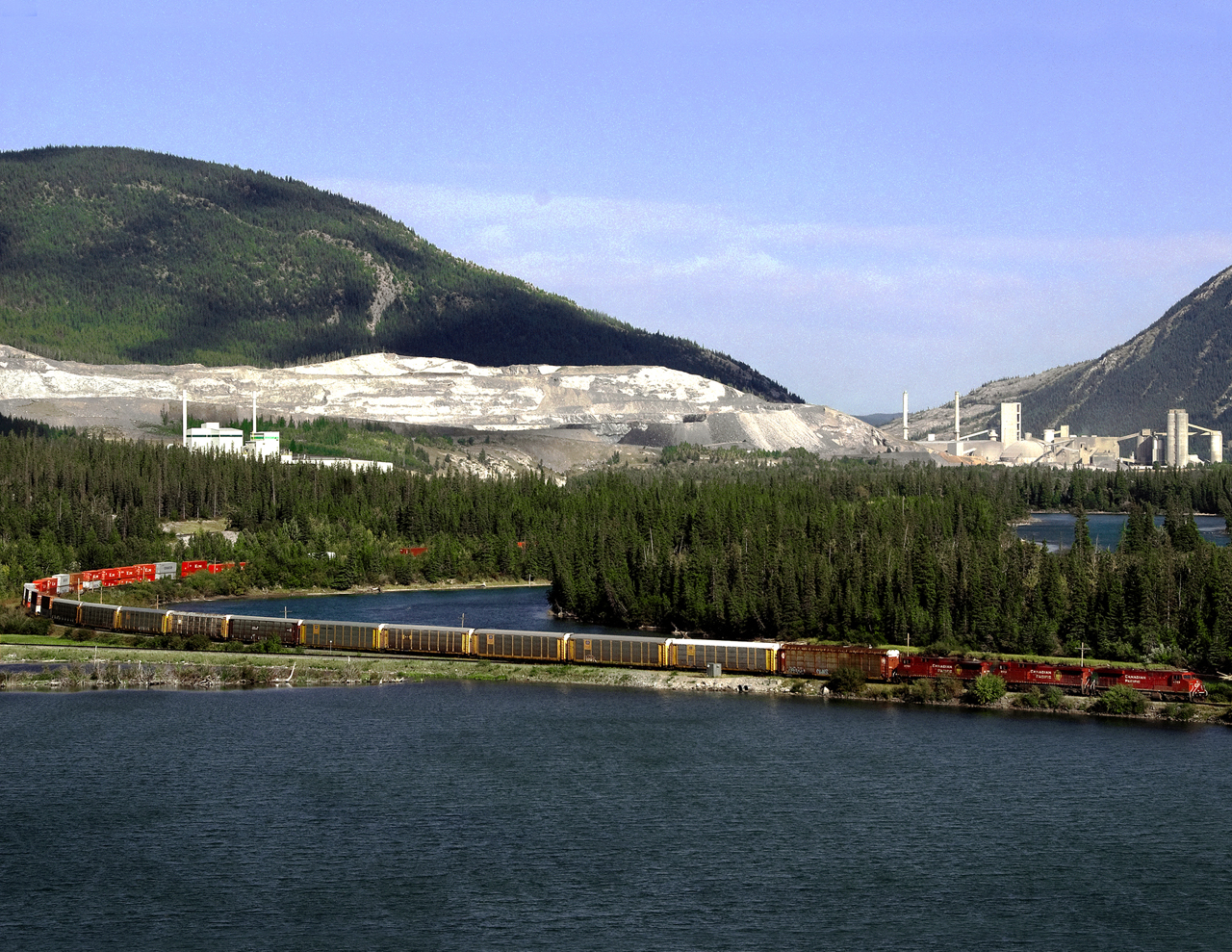 Westbound autos/containers train 115 passes Gap Lake in the Bow River valley. LaFarge's Exshaw cement plant lies behind.