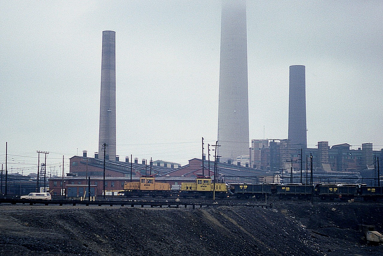 Back in the days of the INCO Electrics, we see motors #123 and 126 rolling past the landmark chimneys of Copper Cliff back in 1985. The barren lands of Greater Sudbury have been considerably cleaned up since these rather dirty times, and green vegetation has returned to the area.
Diesels were introduced to INCO and electrification ended in 2000. Most units scrapped, inc the two shown.
Motor #123 and 126 are both 85T units, built in 1942 and 1950 respectively and purchased for operation in the sprawling Sudbury area complex in 1971 from Kennecott Copper.