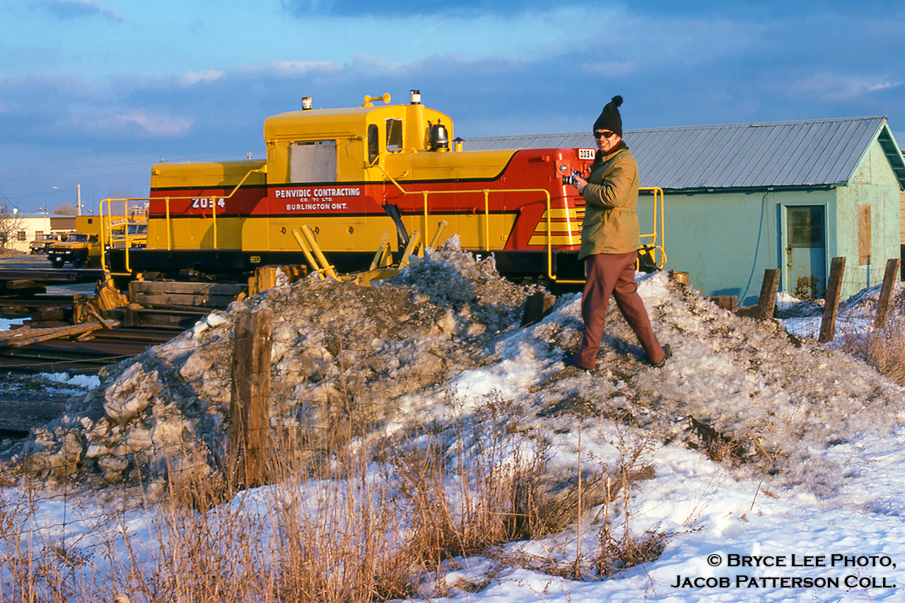 Now who might this be? This individual tagged along with Bryce Lee on a chilly February afternoon in 1977 to check out Penvidic Contracting's ex-CPR 44-ton CLC D-T-C 44H44A1 switcher in a sharp company paint scheme. Note the scheme design is based off the CP maroon and grey it once wore. Built by Canadian Locomotive Co. of Kingston and outshopped on April 29, 1960 as CPR 19, the locomotive was one of 14 D-T-C (Diesel Torque Converter) locomotives, also called DT2s, designed for light switching duties and to be operated without a fireman (weighing less than 45 tons). After it's 12-year career with the CPR, this unit was sold to Penvidic Contracting of Burlington in November, 1972, where it would be involved in a deadly accident while working on the construction of Hydro One's Trafalgar Substation line, branching south off the Galt Sub at the east end of Hornby yard. During the workmen's coffee break on March 14, 1978, the locomotive ran away down a small grade, smashing into cargo vans positioned close to the tracks near where the workers stood. 3 perished, including the engineer who had attempted to board and stop the movement. After an inquest into the incident, the locomotive would be sold in April, 1980 to Aquitaine Petroleum and sent to work at their gas plant in Ram River, Alberta (which also rostered Canada's only ALCO C415s) as their 2034, later to it's original number of 19, and relettered for Aquitaine's new owner, Canterra Energy. It's final operation would be with Skibstead in Rosebud, Alberta before being retired in September, 1987 and put on display in Champion Park, Okotoks, Alberta.Bryce's shot once he climbed up the snowbank.More DT2s:

Dan Dell'Unto Collection: Dryden, Ontario.
Bill Hooper: Canal Flats, British Columbia.
Bill Thomson: Goderich, Ontario.
Bill Thomson: Tottenham, Ontario.Bryce Lee Photo; Jacob Patterson Collection slide.