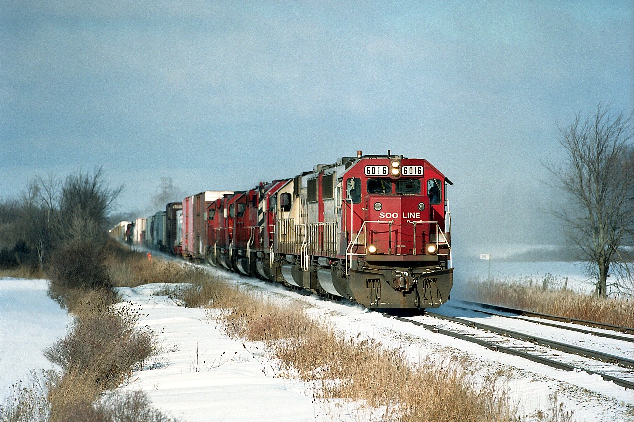 Cold and windy day; at least sunny most of the time, as an eastbound freight kicks up a dusting of the white stuff as it barrels along heading for Welland. Note mile marker 38 on the right. Impressive lineup of power on the head end includes: SOO 6016, 6003, STLH 5615, CP 5585, 5541 and 5825.