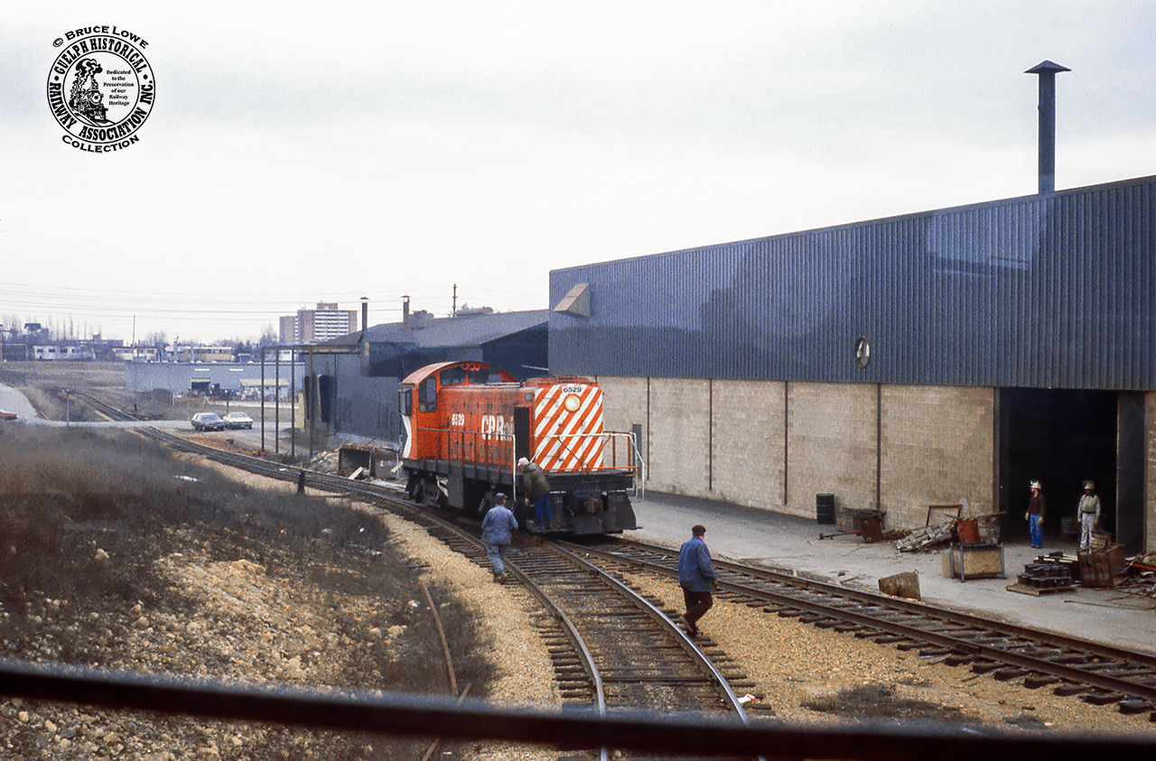 Taken from the cab of SW8 7170 (GMD, 1951) on the CN Guelph Yard job, a meet is seen with the CP yard job, led by S3 6529 (MLW, 1955). CP is seen proceeding up the grade from Dawson Road towards Maple Leaf Mills, today Traxxside Transloading. At this time the facility only had one spur on the property.  CN 7170 sits clear of the South Industrial main on the lead to National Standard; today Traxx 5 and 6; the Traxxside glass tracks. Both locomotives would be off the roster by 1986. The facility pictured behind is Court Galvanizing Ltd.