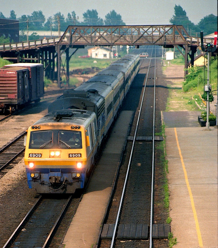 A westbound VIA train of conventional coaches speeds through Woodstock station led by LRC 6909 and a B unit.  There are boxcars in the small yard, a semaphore train order signal and the old Bay Street bridge.  The station is still there, but everything else has gone.