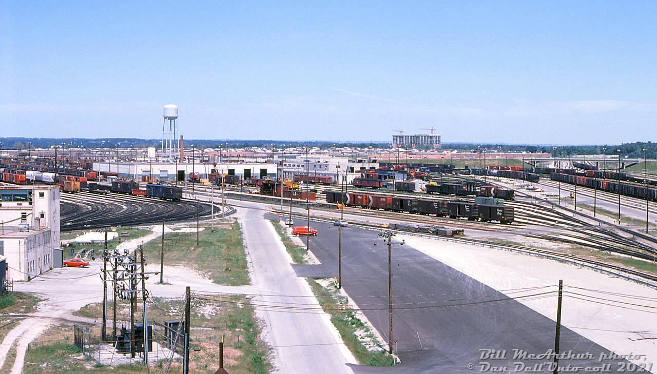 Here's a sunny afternoon view of CP's Toronto Yard (a.k.a. Agincourt Yard) on July 2nd 1978, as viewed looking east from the General Yard Office. Visible are the Car Shops and repair yards, Diesel Shop and servicing areas (and its water tower), and the West Receiving and East Recieving yards to the right. Markham Road is visible in the distance, and the two mainline tracks of the Belleville Sub pass under it on the far right side of the underpass. On the left is the Retarder tower, and part of the sprawling Classification Yard tracks. 

Scarborough's suburban housing sprawl is present in the distance, swallowing up land that was once rural farmland and fields that existed all around the yard when it was originally built "out in the boonies" in the early 60's.

The Car Shop yard is dominated by "home road" CP cars, the most visible are tonnes classic 40' boxcars in stepped lettering (1950's), script lettering (1960's) and action red (1970's). Baggage cars in OCS service and  vans (cabooses) are also present.

Bill McArthur photo, Dan Dell'Unto collection slide.