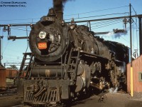 Canadian National Railways U-3-a 4-8-4 6300 rests over the ash pit on the servicing tracks at the Sarnia roundhouse during the later years of the steam era.  Transferred north from the states during December 1941 and based out of Mimico, the locomotive originally worked for CNR’s American subsidiary, Grand Trunk Western, and had been built by ALCO in July 1927 as part of a twelve unit order (6300 – 6311).  It would later be scrapped in April 1959.  Note the lack of ‘Grand Trunk’ lettering on the number plate, which had been removed after transfer to CNR.  The interesting contraption overhead straddling three of the tracks is possibly used in connection with the ash pit, perhaps to aid in emptying into OCS gondolas usually kept on the centre track.<br><br>At far left the catenary of the <a href=http://www.railpictures.ca/?attachment_id=27955>St. Clair Tunnel Company</a> electric operations can be seen.  The St. Clair Tunnel Company, a Grand Trunk Railway and later CNR subsidiary, began operations in 1891 with the <a href=http://www.trainweb.org/oldtimetrains/photos/cnr_electric/StCTCo_1308.jpg>completion of its namesake tunnel beneath the St. Clair River</a> connecting to Grand Trunk Western Railroad operations at Port Huron, Michigan, replacing car float service.  Initial operations began with <a href=http://www.rrpicturearchives.net/showPicture.aspx?id=4354468>four Baldwin 0-10-0 Decapod camelback tank engines</a> numbered 598 – 601, which were later modified with <a href=http://www.trainweb.org/oldtimetrains/photos/cnr_electric/StCTCo_1304.jpg>the addition of tenders in 1898.</a>  Electric operations began in 1908 with six boxcab locomotives from Baldwin – Westinghouse.  After half a century of electric service, the wires were deenergized on September 28, 1958 with the advent of diesels and all electrics were scrapped within the year.<br><br>By the latter half of the twentieth century trains had outgrown the size of the St. Clair Tunnel, leading to the construction of a new tunnel, the <a href=http://www.railpictures.ca/?attachment_id=3944>Paul M. Tellier,</a> which had been <a href=http://www.railpictures.ca/?attachment_id=23890>constructed throughout 1993 – 1994</a> officially opening for service April 5, 1995.<br><br>More U-3-a in Canada:<br><a href=http://www.railpictures.ca/?attachment_id=13305>6301 at Clarkson by bill Thomson, 1953.</a><br><a href=http://www.railpictures.ca/?attachment_id=18760>6305 at Niagara Falls by Del Rosamond, 1959.</a><br><br><i>Original Photographer Unknown, Al Chione Duplicate, Jacob Patterson Collection slide.</i>