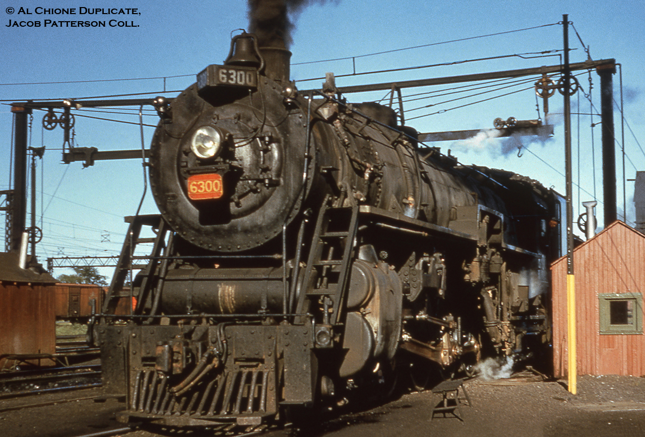Canadian National Railways U-3-a 4-8-4 6300 rests over the ash pit on the servicing tracks at the Sarnia roundhouse during the later years of the steam era.  Transferred north from the states during December 1941 and based out of Mimico, the locomotive originally worked for CNR’s American subsidiary, Grand Trunk Western, and had been built by ALCO in July 1927 as part of a twelve unit order (6300 – 6311).  It would later be scrapped in April 1959.  Note the lack of ‘Grand Trunk’ lettering on the number plate, which had been removed after transfer to CNR.  The interesting contraption overhead straddling three of the tracks is possibly used in connection with the ash pit, perhaps to aid in emptying into OCS gondolas usually kept on the centre track.At far left the catenary of the St. Clair Tunnel Company electric operations can be seen.  The St. Clair Tunnel Company, a Grand Trunk Railway and later CNR subsidiary, began operations in 1891 with the completion of its namesake tunnel beneath the St. Clair River connecting to Grand Trunk Western Railroad operations at Port Huron, Michigan, replacing car float service.  Initial operations began with four Baldwin 0-10-0 Decapod camelback tank engines numbered 598 – 601, which were later modified with the addition of tenders in 1898.  Electric operations began in 1908 with six boxcab locomotives from Baldwin – Westinghouse.  After half a century of electric service, the wires were deenergized on September 28, 1958 with the advent of diesels and all electrics were scrapped within the year.By the latter half of the twentieth century trains had outgrown the size of the St. Clair Tunnel, leading to the construction of a new tunnel, the Paul M. Tellier, which had been constructed throughout 1993 – 1994 officially opening for service April 5, 1995.More U-3-a in Canada:6301 at Clarkson by bill Thomson, 1953.6305 at Niagara Falls by Del Rosamond, 1959.Original Photographer Unknown, Al Chione Duplicate, Jacob Patterson Collection slide.