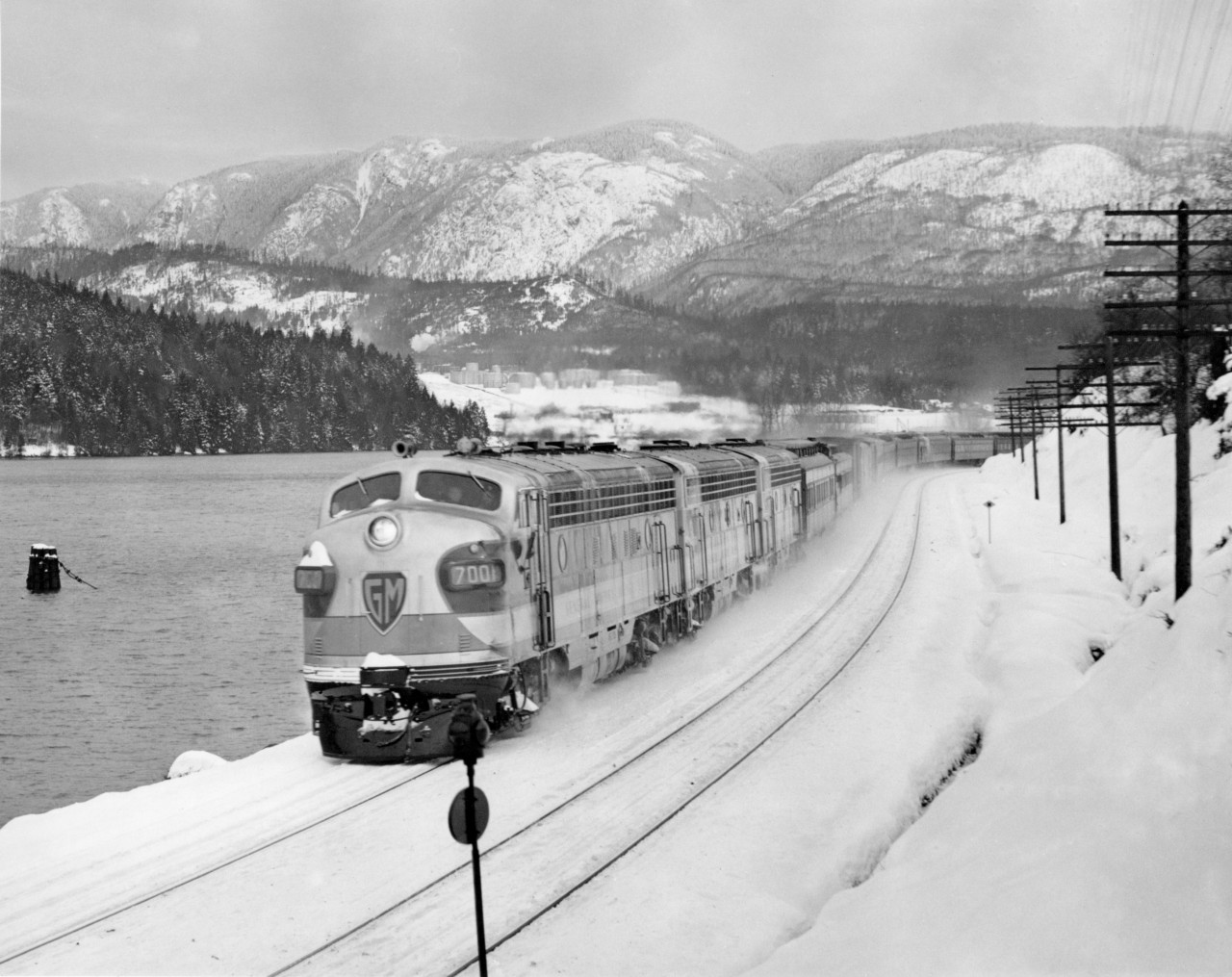 CPR's train no. 7, the first one into Vancouver powered by 4500 H.P. diesel electric on loan to the CPR from General Motors.  The train was almost three hours late.  Consisted of 13 cars and arrived in Vancouver on Friday the 13'th of January, 1950.  Burrard Inlet west of Port Moody by 1.5 miles.

"TEST RUN DOWN FRASER VALLEY   CPR diesel Opens New Rail Era"   By A. J. Darrymple 
  Out of this world!  That's the three-unit royal blue, silver and gold General Motors diesel that pulled CPR's crack transcontinental "Dominion" into Vancouver at 10:50 a.m. today in a history-making test run heralding a new era in transportation developement in Canada.
  Imagine yourself at the head of a power plant 154 feet eight inches in length, with total weight of 718,000 pounds, lifting a string of 13 steel coaches, 1190 tons, up the grades and rolling around the heavy curvatures at 55 miles an hour, and you realize the thrill of railroading.
  Although these diesels have been undergoing exhaustive tests in Ontario, this test run really started from Winnipeg for the 1473 miles to Vancouver under sever winter conditions.
  Reporters, radio newscasters and photographers boarded the train at North Bend before dawn today for the run over the last lap into Vancouver.
  A blizzard roared down the Fraser canyon; and officials of General Electric and CPR checked the operation of the three units under sub-zero conditions.
  In the big, clean, roomy, clear-vision cab, the experts scanned the operational devices and speedmeter as the powerful engines sang their song of high efficiency.
  Veteran Engineer Alfred Fahey handled the throttle with Fireman M. J. Marshall on the left-hand side.  While the window-wipers beat metronome time to the humming wheels, the crew, flanked by a battery of maintenence men and operating instructors, leaned forward peering through the snowy mists.
  Down through Yale and then the frosty Fraser Valley, rolling toward Deroche, passing the stations in a swirl of snow, all intent upon the flashing signals.
  A pin point of green.
  "Clear board," sang the engineer.
  "Clear board," corroborated his mate.
  On through Dewdney and Hatzic, passing the eastbound Huntingdon mixed train at 9:37, and then reaching out for the curving course around Burrard inlet, and through the yards and waving crowds at the station.
  "A fine trip," said D. M. Campbell, General Motors application engineer, "but we would have preferred the test under more rigorous conditions; 15 below was the worst we went through."
  Regardless of the weather, CPR chalked up another first; the first of such power units to be used in transcontinental passenger train through the Rockies; recalling another first, that day, May 23, 1887, when old No. 374 pulled the first passenger train into Vancouver.
  Now the trend is distinctly from time - honored steam to diesel; lower fuel, maintenance and labor costs; improved schedules through reduction of service steops.  Example:  Winnipeg - Vancouver run steam locomotives make 32 stops for water; diesels 10.
  For the passenger:  Smoother stops and starting and a more restful ride; and for the romantic - minded rail-fan, a sleek machine in brilliant color designs, running swiftly and purring like an electric clock.
  CPR officials aboard were T. F. Donald, Winnipeg, assistant superintendent of motive power; G. H. Nowell, Vancouver district master mechanic;  Al Cowburn, assistant superintendent, North Bend; W. Sinclair, Montreal, diesel equipment engineer;  B. W. Woodland, Winnipeg, internal combustion equipment superinvisor; Ernest Hanson, Vancouver, diesel supervisor for Pacific region.
  General Motors officials:  H. D. Dana, assistant general servicee manager;  R. E. Hunter, sales director;  Don Campbell, engineer;  Mel Johnson, service and maintenance engineer;  Frank Switzer, Ed Park, E. Formento, operating instructors.