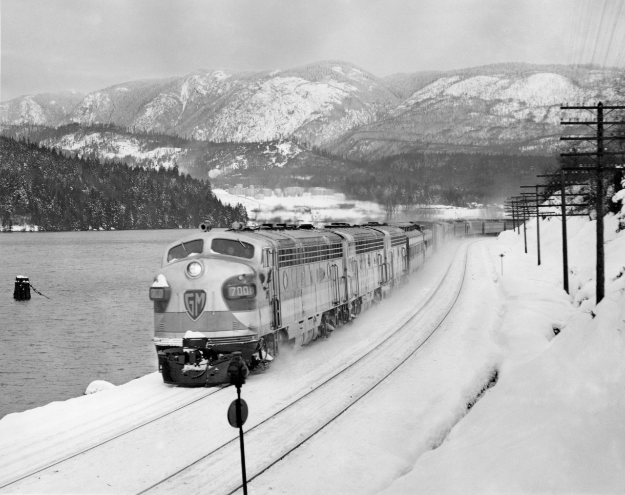 CPR's train no. 7, the first one into Vancouver powered by 4500 H.P. diesel electric on loan to the CPR from General Motors.  The train was almost three hours late.  Consisted of 13 cars and arrived in Vancouver on Friday the 13'th of January, 1950.  Burrard Inlet west of Port Moody by 1.5 miles.

"TEST RUN DOWN FRASER VALLEY   CPR diesel Opens New Rail Era"   By A. J. Darrymple 
  Out of this world!  That's the three-unit royal blue, silver and gold General Motors diesel that pulled CPR's crack transcontinental "Dominion" into Vancouver at 10:50 a.m. today in a history-making test run heralding a new era in transportation developement in Canada.
  Imagine yourself at the head of a power plant 154 feet eight inches in length, with total weight of 718,000 pounds, lifting a string of 13 steel coaches, 1190 tons, up the grades and rolling around the heavy curvatures at 55 miles an hour, and you realize the thrill of railroading.
  Although these diesels have been undergoing exhaustive tests in Ontario, this test run really started from Winnipeg for the 1473 miles to Vancouver under sever winter conditions.
  Reporters, radio newscasters and photographers boarded the train at North Bend before dawn today for the run over the last lap into Vancouver.
  A blizzard roared down the Fraser canyon; and officials of General Electric and CPR checked the operation of the three units under sub-zero conditions.
  In the big, clean, roomy, clear-vision cab, the experts scanned the operational devices and speedmeter as the powerful engines sang their song of high efficiency.
  Veteran Engineer Alfred Fahey handled the throttle with Fireman M. J. Marshall on the left-hand side.  While the window-wipers beat metronome time to the humming wheels, the crew, flanked by a battery of maintenence men and operating instructors, leaned forward peering through the snowy mists.
  Down through Yale and then the frosty Fraser Valley, rolling toward Deroche, passing the stations in a swirl of snow, all intent upon the flashing signals.
  A pin point of green.
  "Clear board," sang the engineer.
  "Clear board," corroborated his mate.
  On through Dewdney and Hatzic, passing the eastbound Huntingdon mixed train at 9:37, and then reaching out for the curving course around Burrard inlet, and through the yards and waving crowds at the station.
  "A fine trip," said D. M. Campbell, General Motors application engineer, "but we would have preferred the test under more rigorous conditions; 15 below was the worst we went through."
  Regardless of the weather, CPR chalked up another first; the first of such power units to be used in transcontinental passenger train through the Rockies; recalling another first, that day, May 23, 1887, when old No. 374 pulled the first passenger train into Vancouver.
  Now the trend is distinctly from time - honored steam to diesel; lower fuel, maintenance and labor costs; improved schedules through reduction of service steops.  Example:  Winnipeg - Vancouver run steam locomotives make 32 stops for water; diesels 10.
  For the passenger:  Smoother stops and starting and a more restful ride; and for the romantic - minded rail-fan, a sleek machine in brilliant color designs, running swiftly and purring like an electric clock.
  CPR officials aboard were T. F. Donald, Winnipeg, assistant superintendent of motive power; G. H. Nowell, Vancouver district master mechanic;  Al Cowburn, assistant superintendent, North Bend; W. Sinclair, Montreal, diesel equipment engineer;  B. W. Woodland, Winnipeg, internal combustion equipment superinvisor; Ernest Hanson, Vancouver, diesel supervisor for Pacific region.
  General Motors officials:  H. D. Dana, assistant general servicee manager;  R. E. Hunter, sales director;  Don Campbell, engineer;  Mel Johnson, service and maintenance engineer;  Frank Switzer, Ed Park, E. Formento, operating instructors.