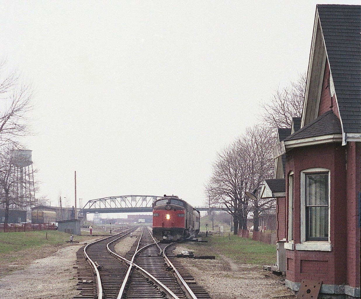 These were the lean years for Amtrak. This train which rattled its way along the CASO from Detroit thru to the International Bridge to Buffalo was given the name "Niagara Rainbow" a rather inventive name for such a clunker.
This train during these lean years ran with old hand-me-down E8A units, as seen in this image. AMTK 407 is photographed on approach to the bridge having just stopped at the passenger station. A few people and a taxi can be seen. On the right in this image is the old B-1 station, moved in 1981 to Oakes Park down on Central Av to keep the old CNR 6218 company. A lot of things have changed in Fort Erie over the years...on the left, the water tower is gone, as is the crew hostel in front. The bridge recently was replaced. In far background the CN shop has been closed and is now a fledgling RR museum. And the Sidekick of the day was along; standing in front of the pole on the left.