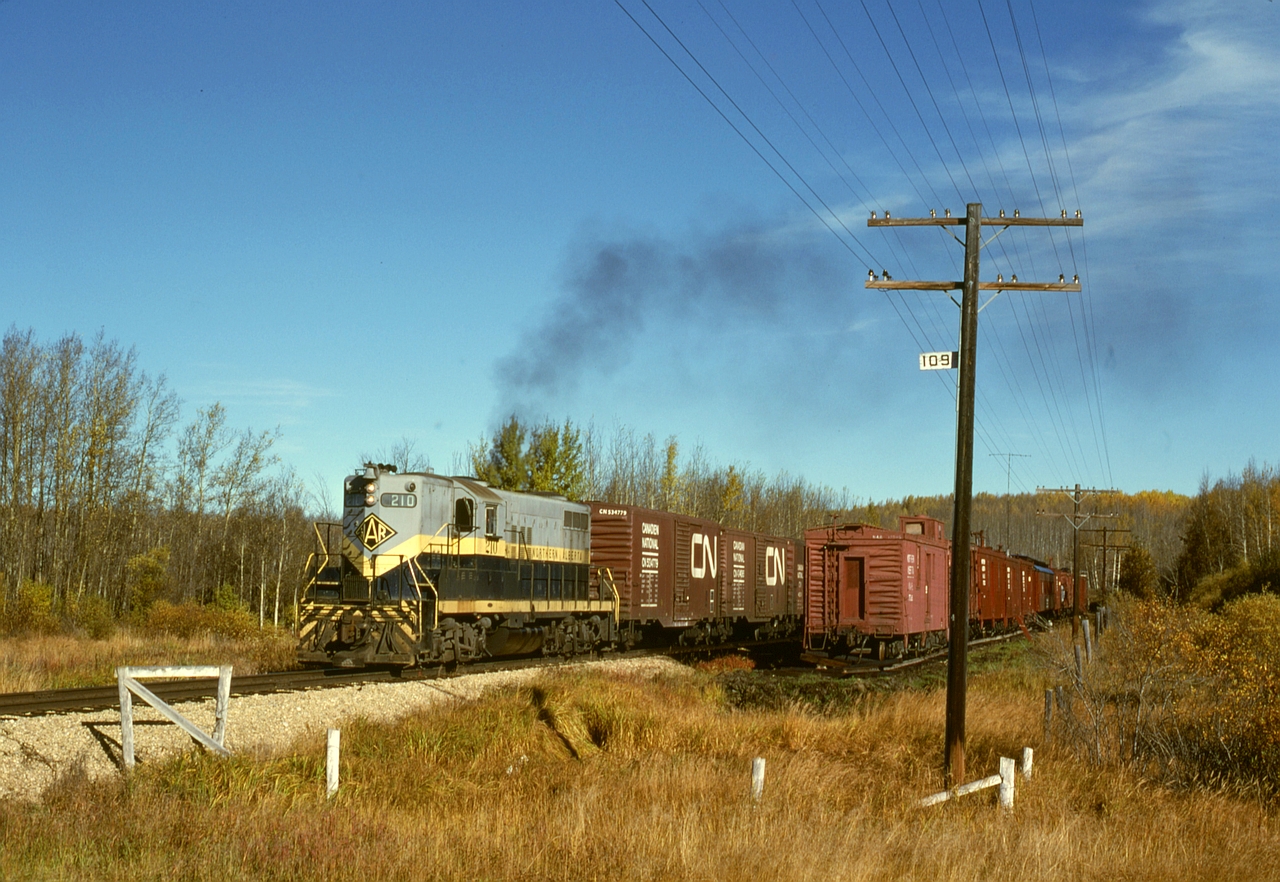 At milepost 109 of the Grande Prairie sub. of the Northern Alberta Railways, highway 43 was crossed, and on the north (timetable west) side there was a spur frequently occupied by bridge and building gang outfit cars for attending to several significant timber trestles over Albright Creek just a bit farther west.  Here is timetable train number 52 with GP9 210 from Dawson Creek, BC, vigorously heading home to Grande Prairie, AB, on Tursday 1977-09-20, just 3.9 km directly east of the BC-Alberta border.  That section of now-CN track is now out of service, and Dawson Creek is served by CN’s former BC Railway lines from the west.