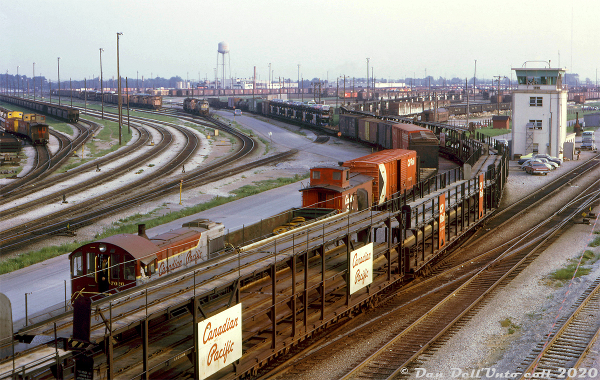 It's a busy summer evening at Toronto (Agincourt) Yard, as viewed from the Finch Avenue overpass at the north end of the yard. A cut of empty open autoracks are being moved around in the foreground as CP S2 7026 switches a mix of cars on the next track over, including a gondola full of freight car wheels in front of its caboose (maybe to/from the car shops). CP 7026 wasn't one of the regular pulldown power units that flat-switched the east end of the hump yard (those were usually a pair of MU'ed MLW S2 or S3 switchers, and only certain numbers were so equipped like 7077+7089, 6563+6564, etc) so 7026 might be working a local, transfer or road job that's just dropping in.

Road freight power in the form of modern MLW and GMD units are visible in the background, with one set running light power (likely heading from the diesel shop in the distance to their train in the yard).

Much of the freight cars in this scene are still in CP's block lettering or script lettering liveries, as new and repainted Action Red cars were just starting to show up. Three vans (in the late 60's "Action Script" colours) are visible on the left near the van shop. On the right is the East Yard Pulldown Tower. Goodies visible in the yard include stock cars (sprayed with white disinfectant lime), covered gondolas, and an old combine passenger car (possibly removed from its trucks and used for storage).

Keith Hansen photo, Dan Dell'Unto collection slide.

More of Toronto-assigned CP S2 7026 around town:

Working the "Old Bruce" service track in West Toronto: http://www.railpictures.ca/?attachment_id=38483

Crossing Strachan Avenue at Parkdale Yard: http://www.railpictures.ca/?attachment_id=35550