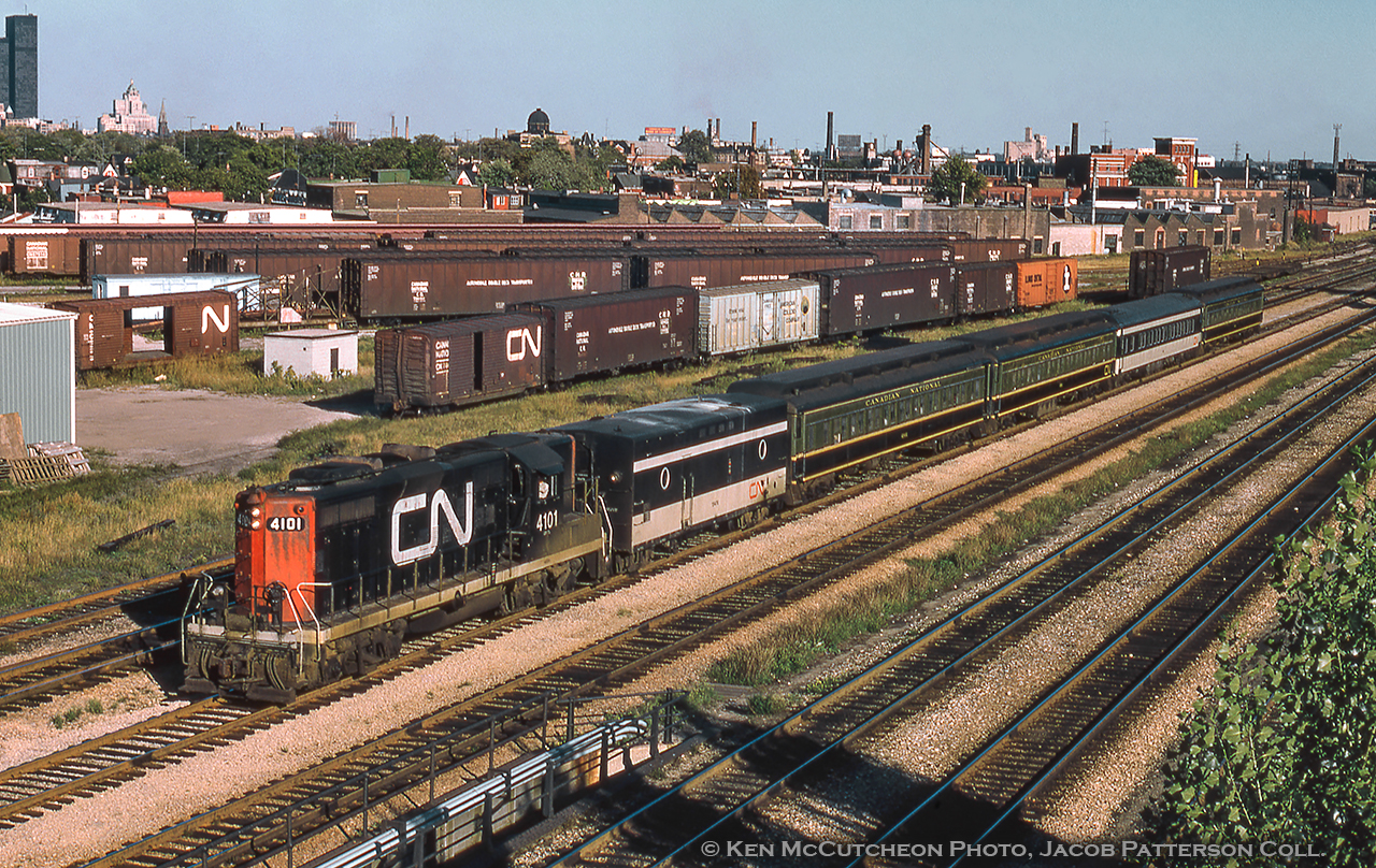 1957-built GP9 4101 hustles train 987, the Guelph commuter, homeward bound on a bright, late summer evening.  Based on the sun angle and time of year it appears to be around 1700h.  While the train is travelling on the CN Weston Sub, the two foreground tracks are the CP Galt Sub leading from West Toronto to Union Station.  A few years later this train will be renumbered 161, likely after the start of GO Transit service to Georgetown in April, 1974 as the consist was shortened at this time.  Lots to see in this image from the heavyweight green and gold clerestory roof cars (the second with a yellow CN noodle in the corner – how common was this?) mixed with steam generator equipment and what appears to be a lightweight CC&F black and white coach.  Looking across the city the all black 56-floor Toronto Dominion Centre, completed 1966, can be seen at far left on the corner of Wellington and Bay Streets, with the Royal York Hotel just to the right (south) on Front Street.  The spire from St. Mary’s Parish can be seen just in front (west) of the hotel, smokestacks from numerous industries dot the landscape, and the red and yellow “Canadian Imperial Bank of Commerce” sign atop the Maple Leaf Mills/Monarch Flour silos tops off the scene on the distant waterfront.A great bit of industry is present at centre in CN’s Parkdale auto yard which is packed with auto carrier boxcars of various types ranging from old 40-footers to the larger CC&F double deck fleet (built 1956 and 1959, totaling 150) with 75 cars at 75 feet in length, and the other 75 at 57 feet.  Labelled “AUTOMOBILE DOUBLE DECK TRANSPORTER,” most of the cars seen still sport their as delivered CNR maple leaf scheme.  As CN’s Car-Go-Rail service diminished in popularity some cars were modified for specific runs (some in Newfoundland, some in Churchill,  seen as recent as 2016!)  Almost half of these cars would go on to Amtrak Auto Train service in 1973, and all remaining cars were retired by 1992.  Note the auto loading ramp a couple tracks above the 4101.*  Today this site is home to a Riverview Produce warehouse, New Choice Excavating & All Type Disposal, as well as condo towers along Dufferin Street.The square tower of the Gladstone Hotel can be seen directly above the lightweight black and white coach on the corner of Queen Street and Gladstone Avenue, right across the street from both the Canadian National and Canadian Pacific Parkdale stations.  Opening in 1889, the hotel originally had an ornate cupola on its tower which was removed during the 1940s.  2004 – 2005 saw the Gladstone undergo a major restoration including its 1903 Victorian elevator, one of the last hand operated ones in Toronto.  Just to the left of the Gladstone another waterfront industry can be seen, Victory Mills Ltd.CN 4101 would be renumbered in 1984 to 4361 (the whole 4100 – 4133 batch renumbered to 4360 – 4393), and later rebuilt in 1991 to 4137.  The unit would be retired in 2000 and sold to the Trans Gabon Railway in Gabon, Africa becoming BB 602.  Current status unknown.A trio of the 40-foot auto boxes sat in Guelph for a number of years alongside an old loading ramp.  These were rumoured to be the last of this type of auto boxcar remaining on CN property, and as such the Guelph Historical Railway Association made attempts to acquire one, CN 740310, built 1948.  About the same time, CN was removing any remaining equipment from the property due to the impending lease to the GEXR in November 1998.  These cars were slated for scrap, however the GHRA had been successful in its requests and the car was to be spared.  Unfortunately, word did not reach the scrap crews in time.  In lieu of this car the GHRA was donated CNR 60337, a 1950 heavy weight baggage car retired from the Hornepayne auxiliary.*Information courtesy of Eric Gagnon.Ken B. McCutcheon Photo, Jacob Patterson Collection Slide.