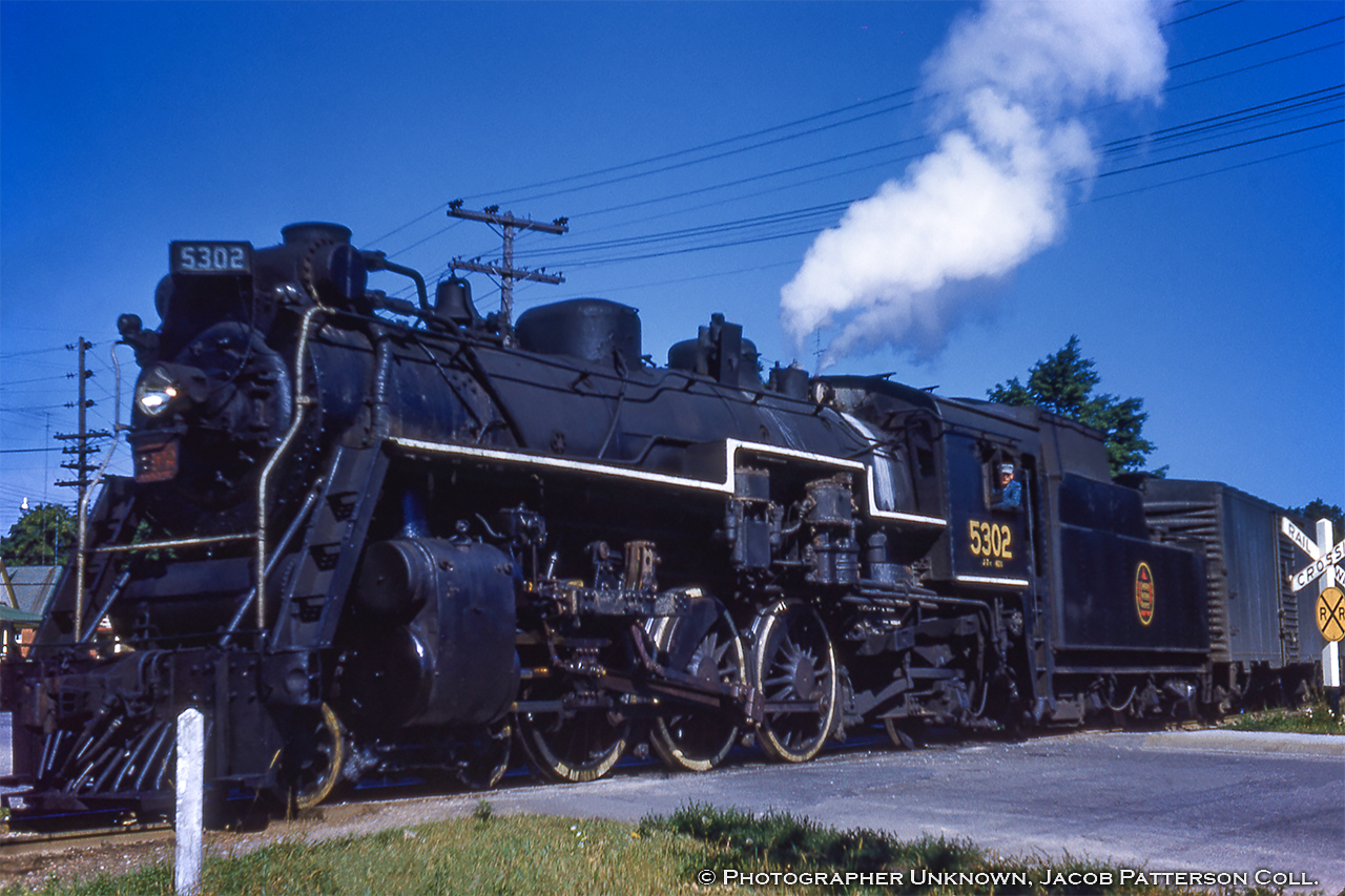 CNR J-7-c Pacific 5302 is seen on the home stretch to its final station stop at Guelph with train 172 crossing Paisley Street at mile 30.47 of the Fergus Subdivision.  The switch just ahead of the locomotive should be lined for the normal position, which at the time routed trains through the Crimea Street wye (Fergus sub main) and onto the Brampton Subdivision.  Crossing Edinburgh Road (mile 49.54) on the Brampton sub, the train will likely take the crossover just to the east onto the south main for the short jaunt to downtown Guelph.  In the background at left it would appear to be the houses at 146 and 148 Edinburgh Road North, though they are in a row of six virtually identical houses.Originating at Owen Sound with a departure time of 0550h (1956 timetable, page 2), 172 would travel south via Palmerston (0800h arrival), before continuing to Guelph arriving at 0940h.  This image was likely snapped just after 0930h.  Based on the timetable the train would return north to Owen sound as train 175, departing Guelph at 1925h, arriving Palmerston at 2045h, and finally due into Owen sound at 2310h.  Both 172 and 175 were daily except Sunday trains.CNR 5302 was built in August 1920 by MLW as part of an order for ten such locomotives (5295 - 5304).  At the  time of this photo it had less than a year to go before meeting the torch in May 1959.Original Photographer Unknown, Jacob Patterson Collection Slide.