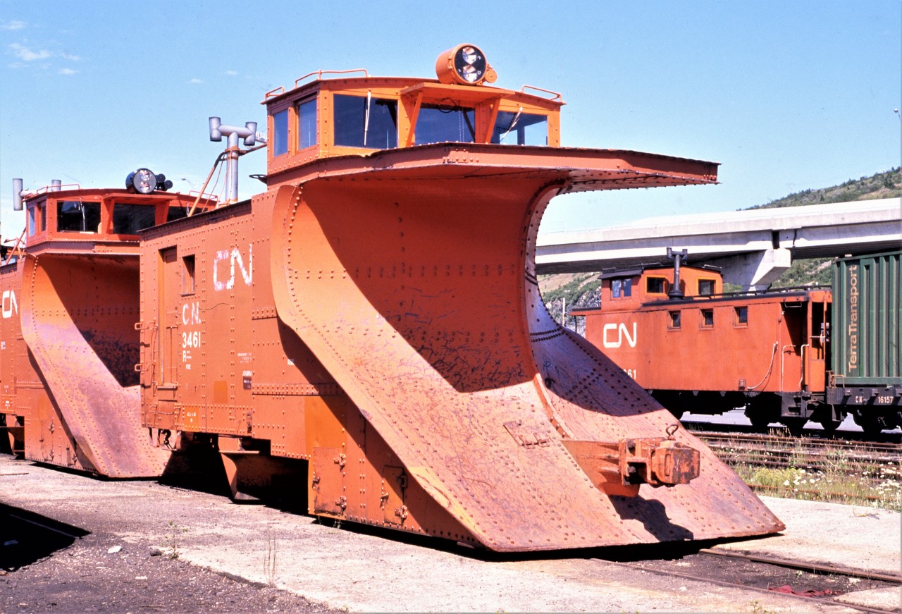 WINTER WARRIOR WAITING. Terra Transport Steel plow No. 3461 waits in the St. John's yard on August 7, 1988 for a winter that never came. Built in a group of twelve (Nos. 3458-3469) by National Steel Car of Hamilton in 1953 after takeover by the CNR (the first group of steel plows for Newfoundland were built by Canadian Car and Foundry in Montreal in 1944, Nos. NR 830-837 to later become CN 3450-3457) it was still on the roster some 33 years later. The 3461 after a lifetime of battling heavy snowdrifts over the Gaff Topsails was living on borrowed time as was the entire narrow gauge operation in Newfoundland. With the impending shutdown to take effect a little over a month later on September 30, it would never again be called to active duty and would see the scrappers blowtorch. Fortunately, first batch sister 3454 is preserved in Bishops Falls as are second batch sisters 3459 in Whitbourne, 3460 in Corner Brook, 3462 in Lewisporte, 3465 in Avondale and 3467 in Port aux Basques. Despite a dwindling of operations, train 203 to the right with caboose 6061 was already marshalled for its 1930hrs departure that evening.