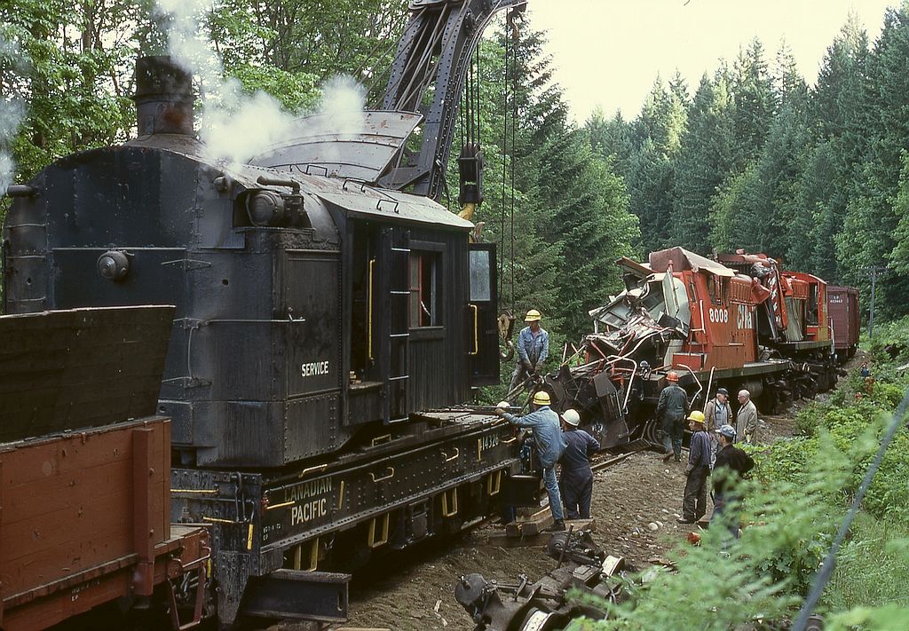 With the Victoria auxiliary on the south end and the Vancouver auxiliary on the north, the cleanup of the day-before head-on at mile 68.3 continued with re-railing the mangled-both-ends lead unit of train 51, 8008﻿, with the crushed cab leaving all involved amazed and thankful the three crew members had survived when trailing unit 8006 had ridden up then over and down the bank to the left, with the behind-the-cab fuel tank of 8008 remarkably intact with no breach.