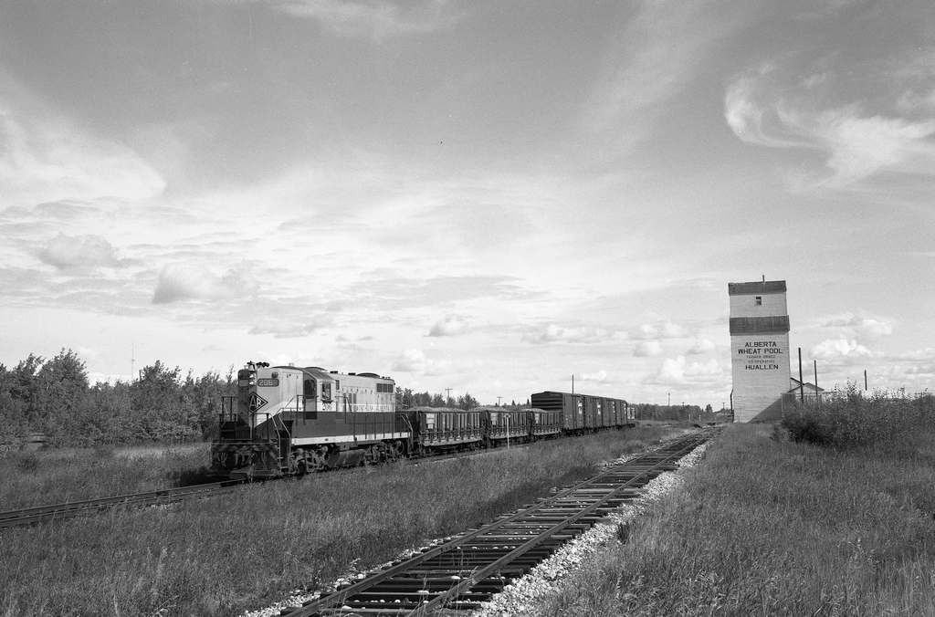 Westward from Grande Prairie, AB, the Northern Alberta Railways fulfilled the earlier name of Edmonton, Dunvegan and British Columbia for this portion with the 88 miles of line to Dawson Creek, BC, and in 1977 were providing thrice weekly wayfreight service with timetable trains No. 51 on Mondays, Wednesdays and Fridays from Grande Prairie and No. 52 on Tuesdays, Thursdays and Saturdays from Dawson Creek.  Motive power was frequently a single GP9, as with this No. 51 engine 206 on Wednesday 1977-09-21 by the lone elevator at Huallen, AB, at mileage 71.1 (measured from Rycroft, with Grande Prairie at 49.7) with three Hart-Otis ballast car loads plus four empty boxcars and caboose 13006.