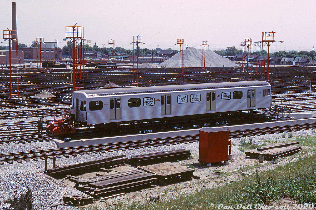 On June 19th 1965, John Bromley was present at the TTC's Greenwood Yard to photograph brand new TTC H1 subway car 5350 being unloaded from its flatcar at the pit track. Shirts appear to be optional on this warm June day as workers ready the car for unloading. At the time the TTC was taking deliveries at Greenwood of new H1 subway cars from Hawker Siddeley in Thunder Bay, ordered for the new crosstown Bloor-Danforth subway line (to open next year, in Feburary 1966) even though the yard was still under construction at the time (note the large gravel piles and stacks of ties in the background).  The H1 subway car bodies were delivered without trucks, so "shop trucks" were slipped under the body at each end. Here, one has already been installed under the rear (blind) end of the car, and one waits on a small trolley to be slipped under the front end on the right. Once the shop trucks are installed, the TTC's red Whiting trackmobile will pull the car off and run it around the bottom of the yard loop track into the shops. The flatcar it's riding on is CP 313003, one of a handful converted from old heavyweight sleeping cars (more info in this photo) to transport new subway cars from MLW and HSC. The pit track the flatcar sits on is standard railway gauge (a siding from CN's Kingston Sub), but the ramp and yard tracks are all TTC gauge (slightly wider, done intentionally to prevent railways from operating over city streetcar tracks),  Two years later, TTC 5350 would be loaded back up onto a CP flatcar for shipment to Thunder Bay for repairs in July 1967, after a sideswipe accident with a Gloucester train damaged the first 1/3rd of right side of the car. It would return and transport the citizens of Toronto underground for over three decades until the H1 fleet was retired en masse in 1997-1999.  John F. Bromley photo, Dan Dell'Unto collection slide.