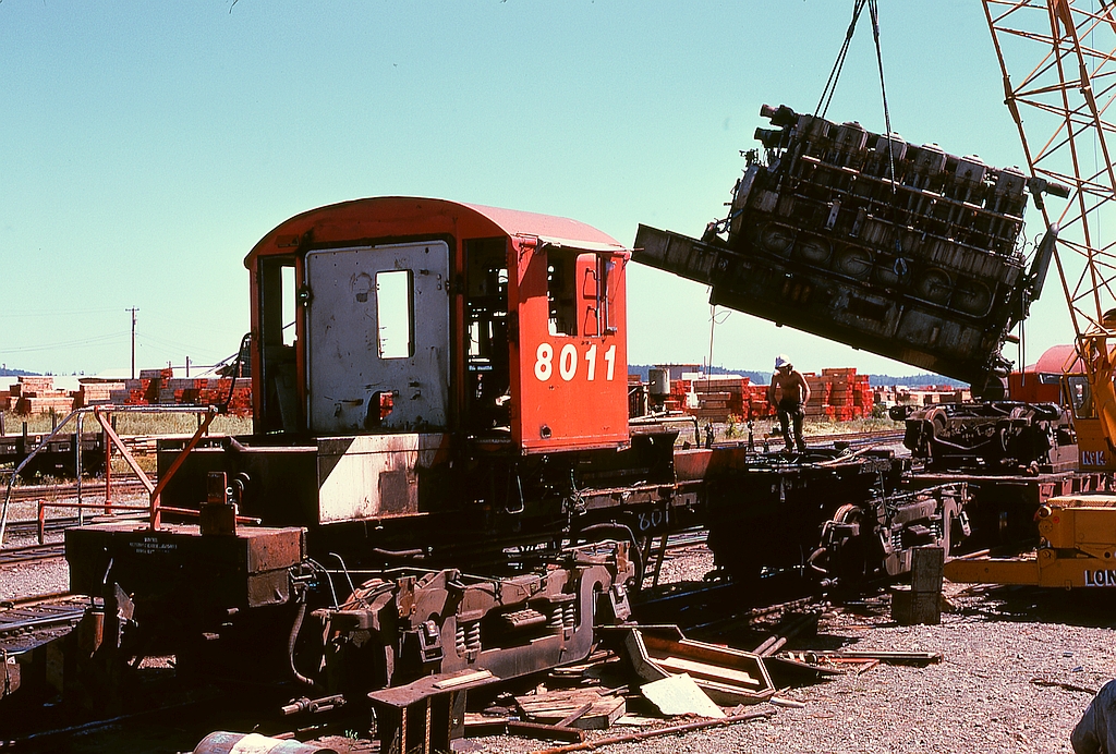 The Baldwin 606SC engine (sans turbo) of 8011 in the air during scrapping at Wellcox yard in Nanaimo on Tuesday 1974-07-30.  One near-comical tension relief during the scrapping was when each engine was first lifted clear.  All shopstaff “knew” there should be a treasure trove of tools lost underneath during earlier years, never to be retrieved unless the engine was removed, but nothing of significance was ever discovered there, much to the prime scavenger’s dismay!