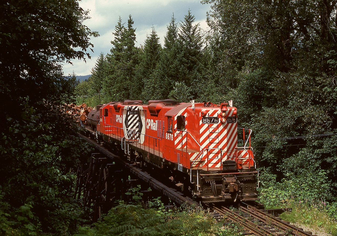 On Vancouver Island, CP’s E&N Division handled logs from the Crown Zellerbach Logging Division reload at Lake Cowichan east 18 miles to Hayward (just north of Duncan) then north 17 miles to Ladysmith and CZ’s log dump.  Here, typically clean GP9s 8676 and 8640 are eastward with a fire-season water car and log loads crossing the awkward-to-photograph (and seldom documented as a result) trestle bridge over Bings Creek at mileage 1.6 west of the junction at Hayward.