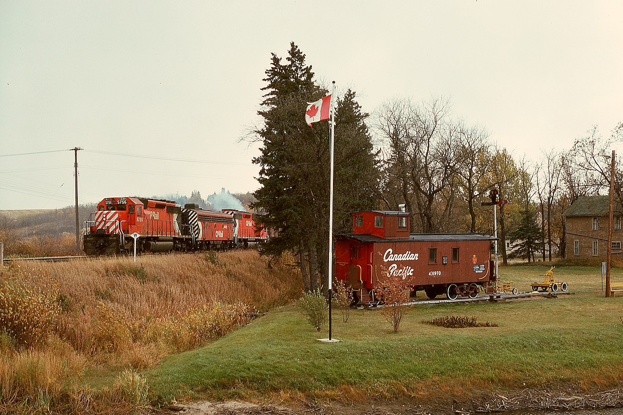 At Minnedosa, MB, where CP’s Minnedosa subdivision from the south joins to the Bredenbury sub. to the west, timetable train No. 977 with SD40-2 5756 and F7B 4439 and SD40 5514 is passing an early railway history display of caboose CP 431970 just before crossing the Little Saskatchewan River then Main Street.  Since then, CP FP7 4038 in maroon/grey with block lettering has joined the caboose in that display.