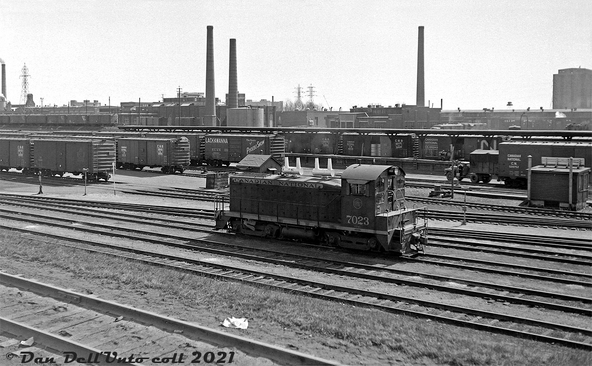 CN SW1200 7023 sits in the east end of Bathurst Street Yard in downtown Toronto, south of Front Street and east of Spadina Avenue, between switching duties in March of 1963. The team tracks and platforms in the background are full with 40' boxcars (the standard freight car of the day for freight, express and LCL shipments) unloading and loading cargo. One can spot a foreign Delaware, Lackawanna & Western (DL&W) "Route of the Phobe Snow" car amid all the CN wafer-logo boxcars, and a new noodle-logo 40' repaint (with yellow doors, initially meant to signify high-class merchandise service before being more commonly associated with CN boxcars in newsprint service).

According to timetables and track diagrams, this part of the yard was the K-200 series of team tracks, often noted as "Lower Yard" (see Robert Farkas' shot here). Hendrie Cartage (a CN affiliated transportation company) operated out of this yard and handled freight and LCL shipments for CN and a variety of customers. Cargo such as food products, newsprint and papers, furniture, textiles (for Toronto's old downtown textile manufacturing industry) was often unloaded from 40' boxcars at the team tracks here, and transloaded onto trucks for local delivery in town, and vice-versa for outbound shipments.

Further east were the K-100 series of express and team tracks next to the CN Express building, that handled express shipments, customs and bonded cargo. Track K-101 was noted as the siding into the Express Building / Bond Shed. K-104 was the "Paper Platform" (outbound express newspaper shipments?), noted customers here include the Globe & Mail, Toronto Daily Star, Toronto Telegram, the Ryerson Press. Hendrie Cartage handled their own print and paper customers at Lower Yard's paper platform (Track K-201).

Auto and truck loads for Car-Go-Rail, auto brokers, select auto dealers, rental companies and manufacturers were noted as handled at the Auto Ramp tracks in the Parkdale area (see here).

The CN Tower parking lot displaced a lot of the K-100 & K-200 team tracks here in the mid-70's, and eventually succumbed to commerical redevelopment (the Metro Toronto Convention Centre, and Royal Bank data centre). The main Bathurst Street yard tracks (K-300 series) were reconfigured in the 1980's for GO Transit train storage, and today are know as GO's Bathurst North Yard. The old CN Express building west of Union Station was renovated into the Skywalk in the 1980's.

The track in the foreground lead up grade to CN's Simcoe Street yard and freight sheds at Front & Simcoe (south of the CP/Dominion Express freight sheds). Both CN and CP had freight sheds here, but CN's were demolished around 1967-68. CP's survived until the late 1970's before being demolished, but some facilities at King St. continued to exist for paper deliveries and team tracks into the 80's.

Background items of interest: CN's prominent Spadina Roundhouse with passenger power such as FPA4 and RS18's visible outside. Further in the background to the left was CP John Street Roundhouse and coach shop, with 40' ice reefers spotted nearby. TTR's Central Heating Plant is just visible off in the distance on the far left. The two smoke stacks in the middle of the photo (between the roundhouses) were for the City of Toronto's water filtration plant at Rees St. The silos on the waterfront at far right were for Maple Leaf Milling (demolished early 80's).

CN 7023: The CN 7000 series were made up of GMD SW9 and SW1200 units, 1200 horsepower "GS-12" switchers not equipped with roadswitcher features such as flexicoil trucks, larger fuel tanks, MU, and full end numberboards like the more common CN 1200- and 1300-series SW1200RS units were. Nevertheless, they along with the lower horsepower SW8 and SW900 units were common across the CN system at various terminals for local and yard switching duties well into the 1980's. Built in September 1956, 7023 later became 7723 (due to the 7000-series GP9RM rebuilds in the 80's that saw larger replacement local and switching power like the GP9's replace smaller, older, lower horsepower units like the SW8/900/9/1200 fleet) and eventually retired and disposed of.

Original photographer unknown, Dan Dell'Unto collection negative (large-format scanned with a DSLR, with some restoration work done).
