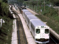 GO Transit cab car 9855 leads a train of five single-level Hawker Siddeley commuter cars eastbound on CN's Oakville Sub towards Brown's Line overpass in southwest Etobicoke, after making its stop at Long Branch GO station. The sun angle suggests this is a late morning train, and timetables indicate GO operated an hourly afternoon service on the Lakeshore Line. An old wooden "Railway Junction, One Mile" triangular sign juts out on the left for eastbound trains approaching Canpa Junction.<br><br>In preparation for the launch of GO Transit's commuter rail service in 1967, it was decided the three old CN station stops serving Long Branch, Dixie Road, and Lakeview were to be consolidated into a new Long Branch GO station at Mile 9.6 of CN's Oakville Sub, located more to the west of the former location (Mile 8.8, by 30th Street) to better connect with the nearby TTC Long Branch streetcar loop. By that time, it doesn't appear there were any station buildings at those old scheduled CN stops, aside from a shelter at Dixie.<br><br><i>Photographer unknown, Dan Dell'Unto collection slide.</i>
